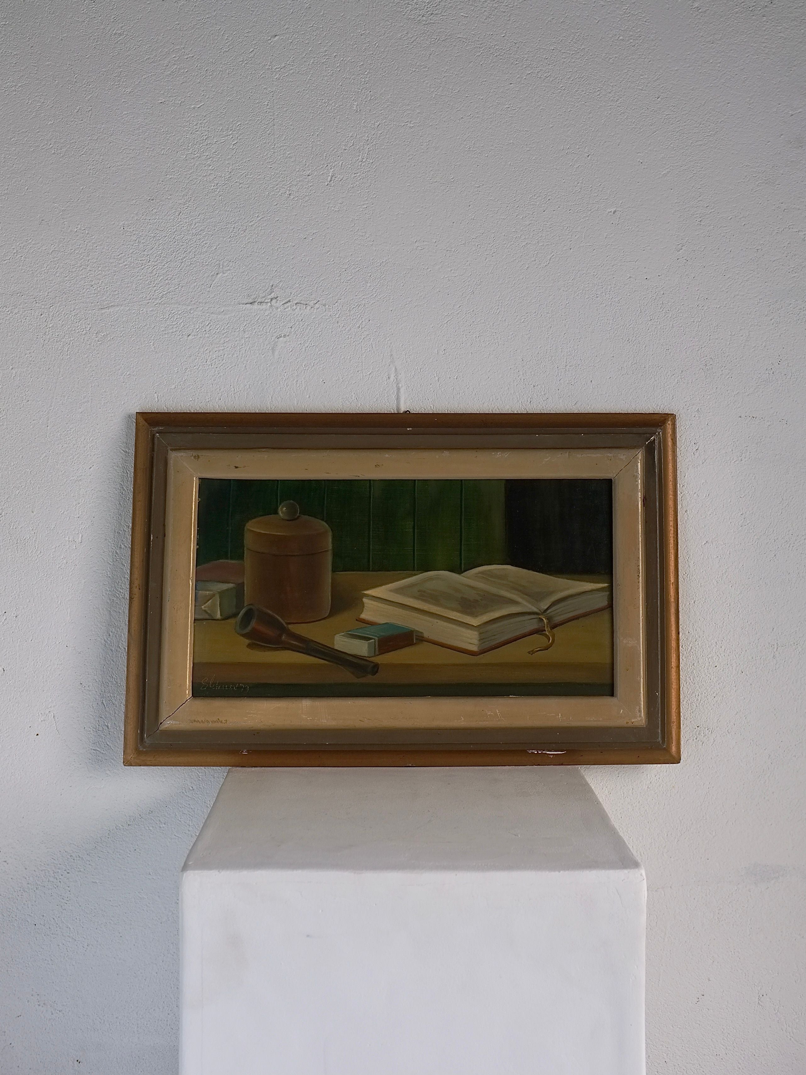 A framed painting of European origin on a pedestal features the Vintage Realism Still Life Oil Painting | 1970s by Veter Vintage, depicting an open book, a wooden mallet, a cylindrical container, and a small square object on a table against a dark green background. The frame is wooden with a metallic inner border and is in vintage condition.