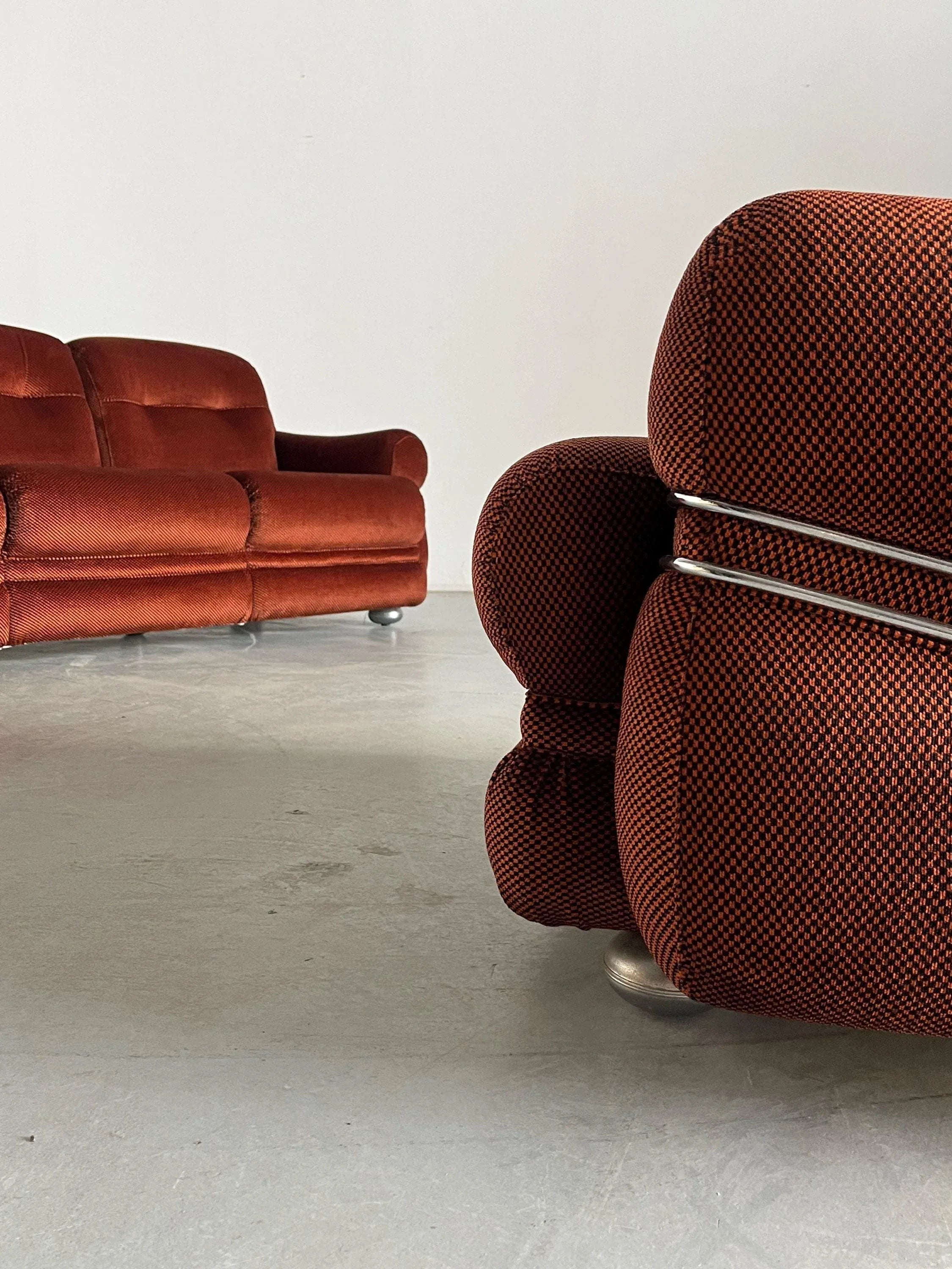 Two Italian Space Age couches from Cherry Cargo with checkered upholstery complement a minimalist room with a light gray floor. One is foregrounded, showcasing its curved armrests and metallic legs, while the other rests subtly in the background, slightly out of focus.