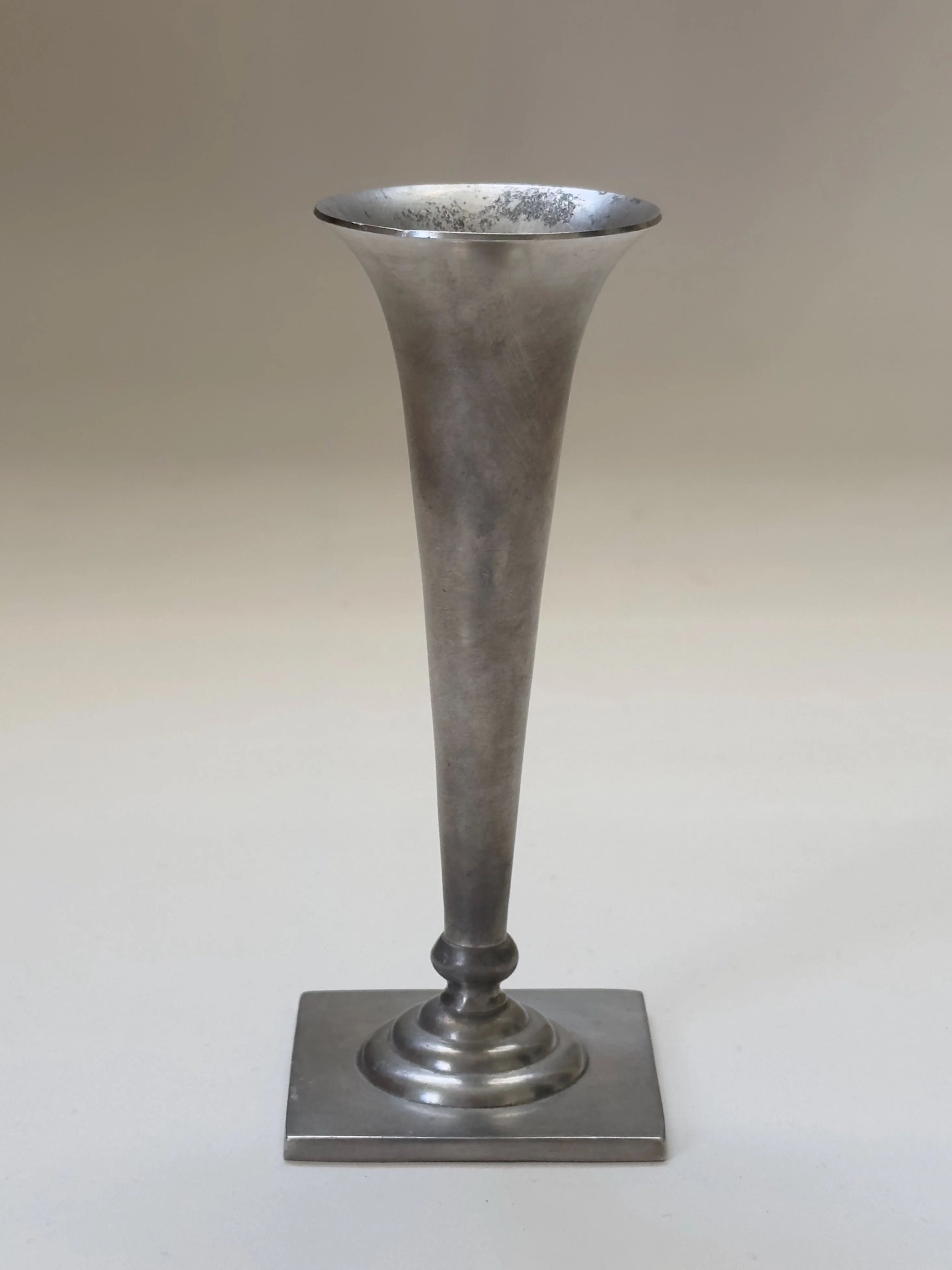 A tall, slender Just Andersen vase by Galerie Storm stands against a plain background. This Art Deco piece, crafted in pewter with a flared top and square base, features a sleek surface that tapers elegantly toward the bottom.