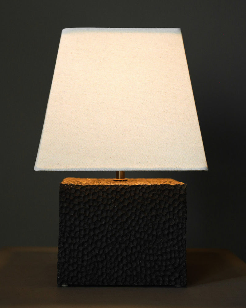 The Akune Ceramic Lamp - Number Five by Studio OSKLO, featuring a textured black rectangular base and wide off-white square shade, is illuminated and casts a warm glow against a dark background.
