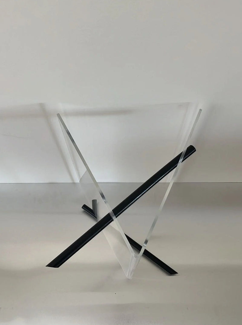 A modern, minimalist Magazine Holder Plexiglass by Lui Art Design made of two intersecting clear acrylic panels forming an 'X' shape, with black accents at the edges where the panels meet. Crafted by CU-RA-TED using premium plexiglass, the stand is set against a plain white background.