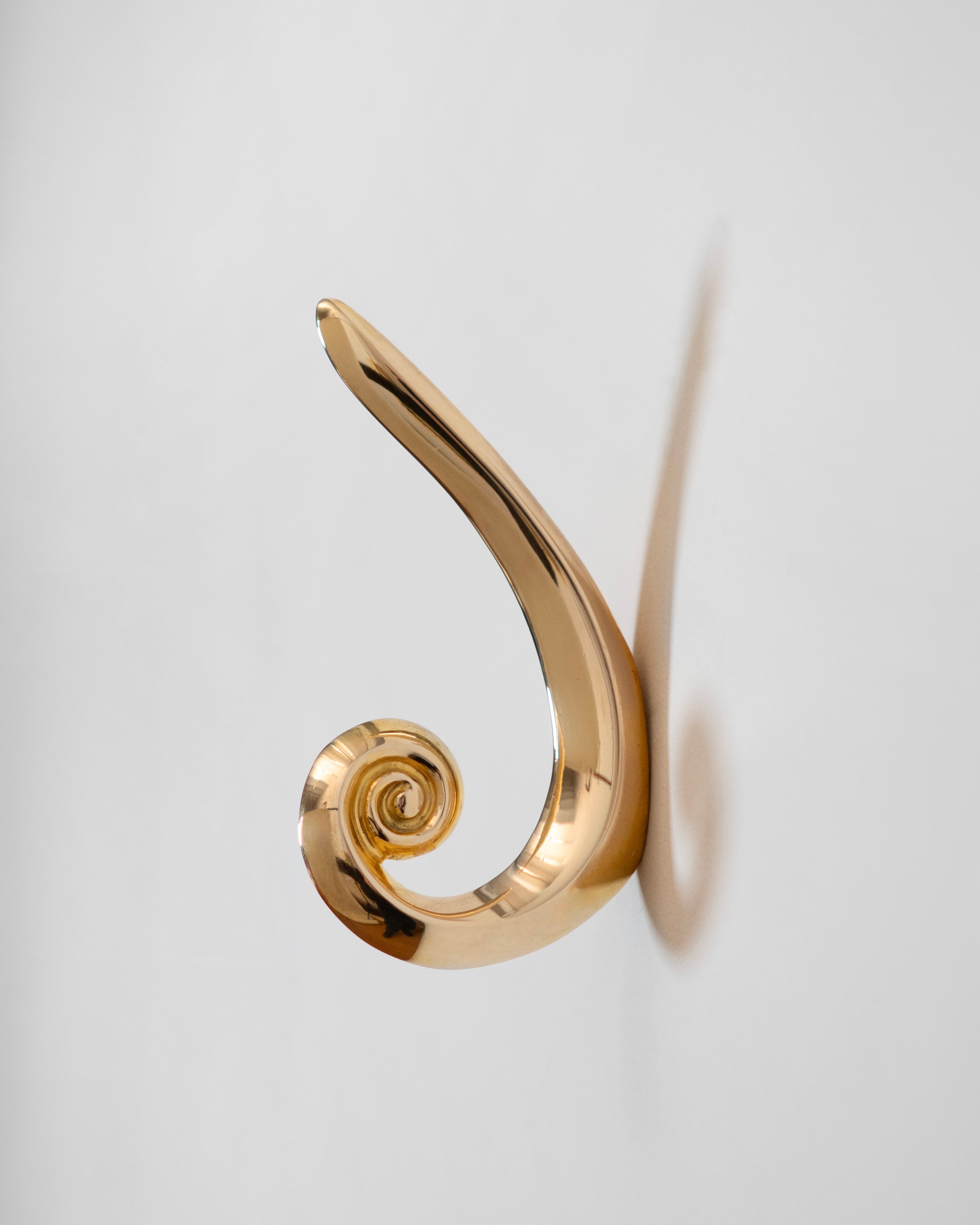 The Noelle Hook - Brass by Hein Studio, featuring a shiny gold finish and abstract spiral design, is mounted on a white wall and casts a soft shadow to the right.