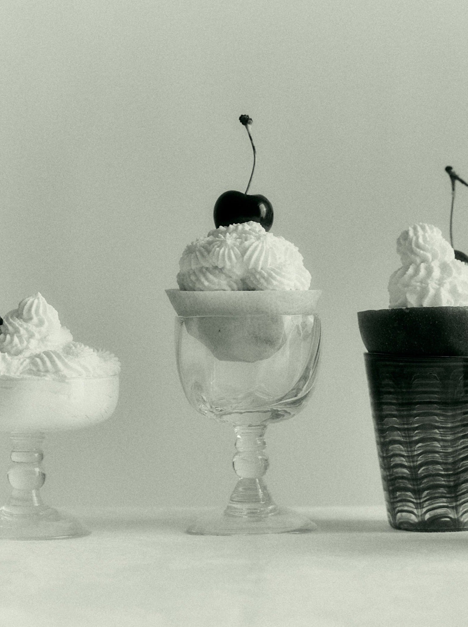 Black and white image of three desserts with whipped cream in Akua Objects Alban Wine Glass dishes, the center one topped with cherries, against a muted background.