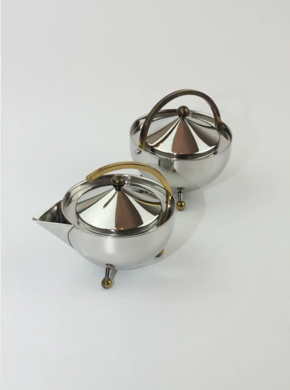 A pair of the Boga Avante Shop Danish Sugar and Creamer Set with gold and brown accents, showcasing elegant Danish design. Both have spherical bodies, narrow spouts, and arched handles. The conical lids feature a finial on top. Each item in the set stands on three small spherical legs against a plain white background.