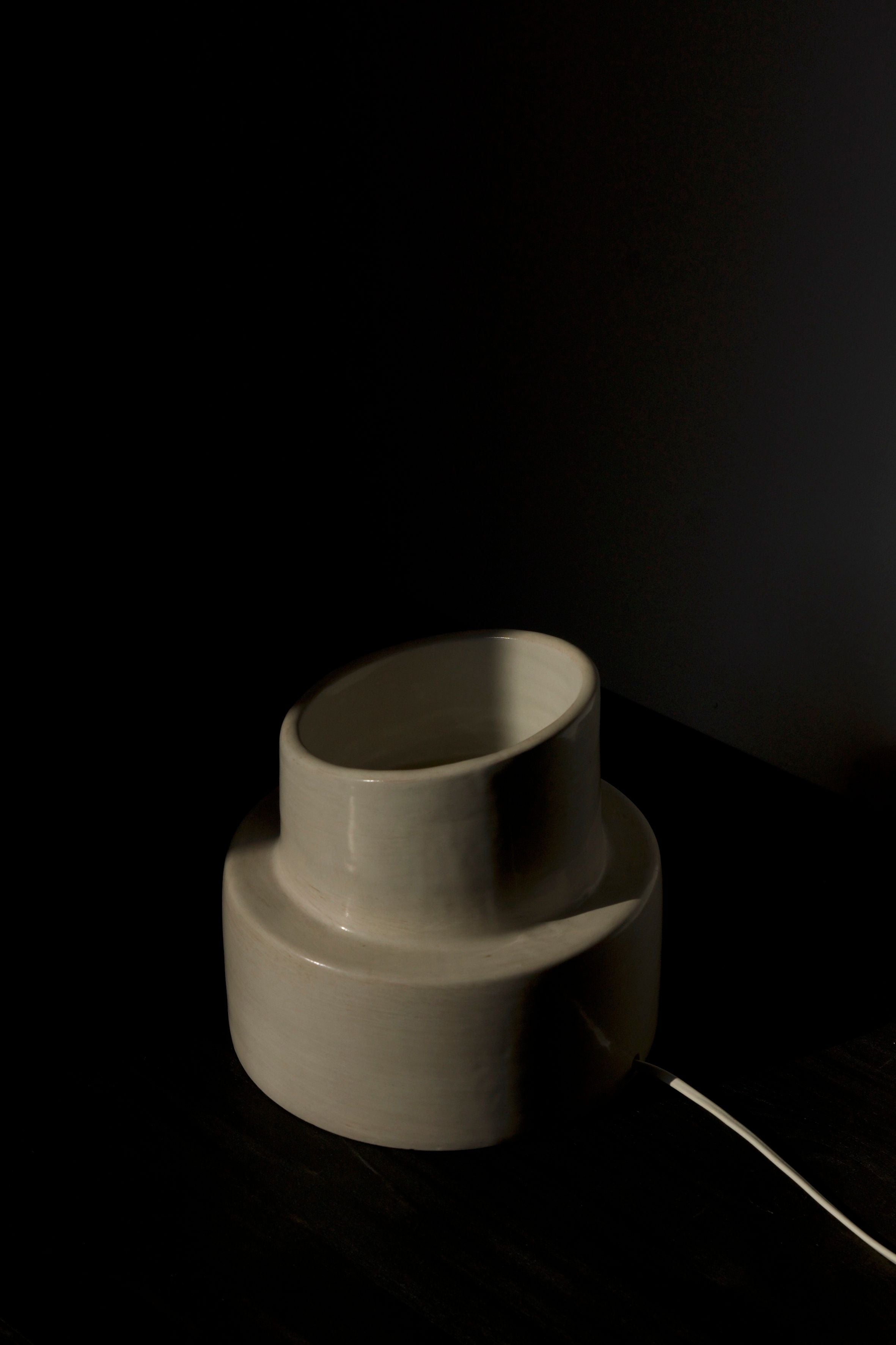 The COSEINCORSO "Pillar" Table Lamp, featuring a rounded, tiered ceramic design, rests on a dark surface. Its white cord extends from the base into shadow, recalling the silhouette of an ancient column.
