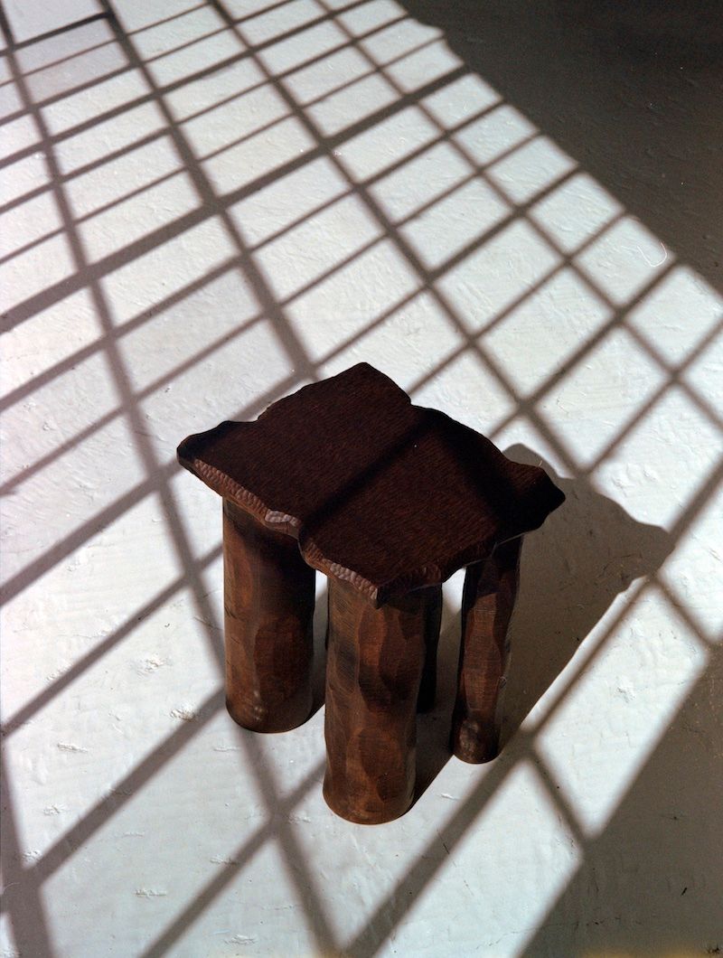 A Mare Side Table by Ombia Studio, crafted from solid white oak with two cylindrical legs, is placed on a sunlit floor. The floor's shadows form a grid pattern, creating a geometric effect.