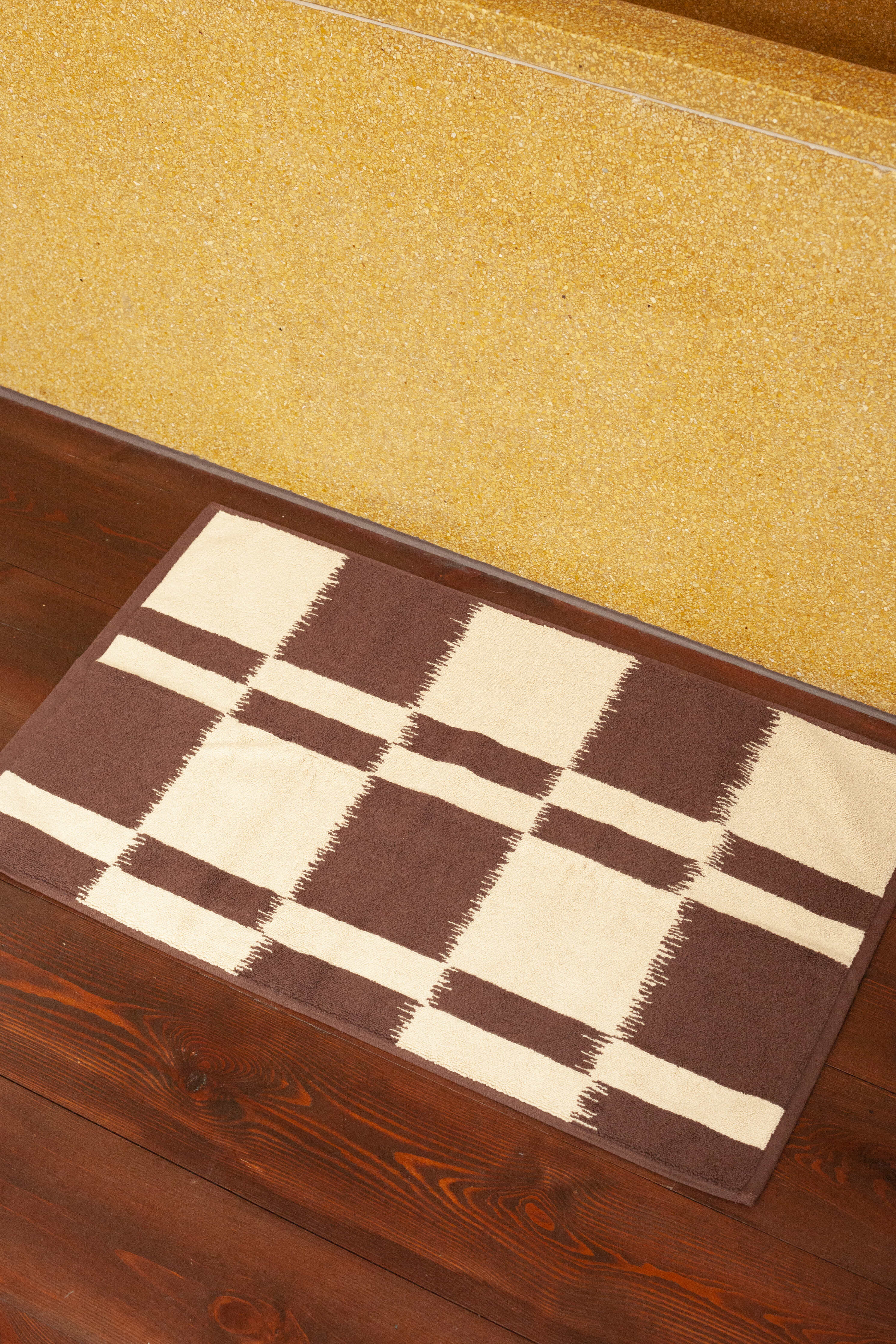 A brown and beige checkered Karin Bath Mat from Autumn Sonata, crafted using Ikat weaving techniques, lies on a dark wooden floor beside a carpeted staircase with a glittery golden surface.