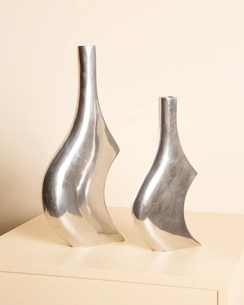 Two abstract Treaptyque vases from the 80's, crafted in silver Italian aluminum, sit on a light surface against a neutral backdrop. These elegant pieces differ in height, with the taller vase gracefully poised on the left.