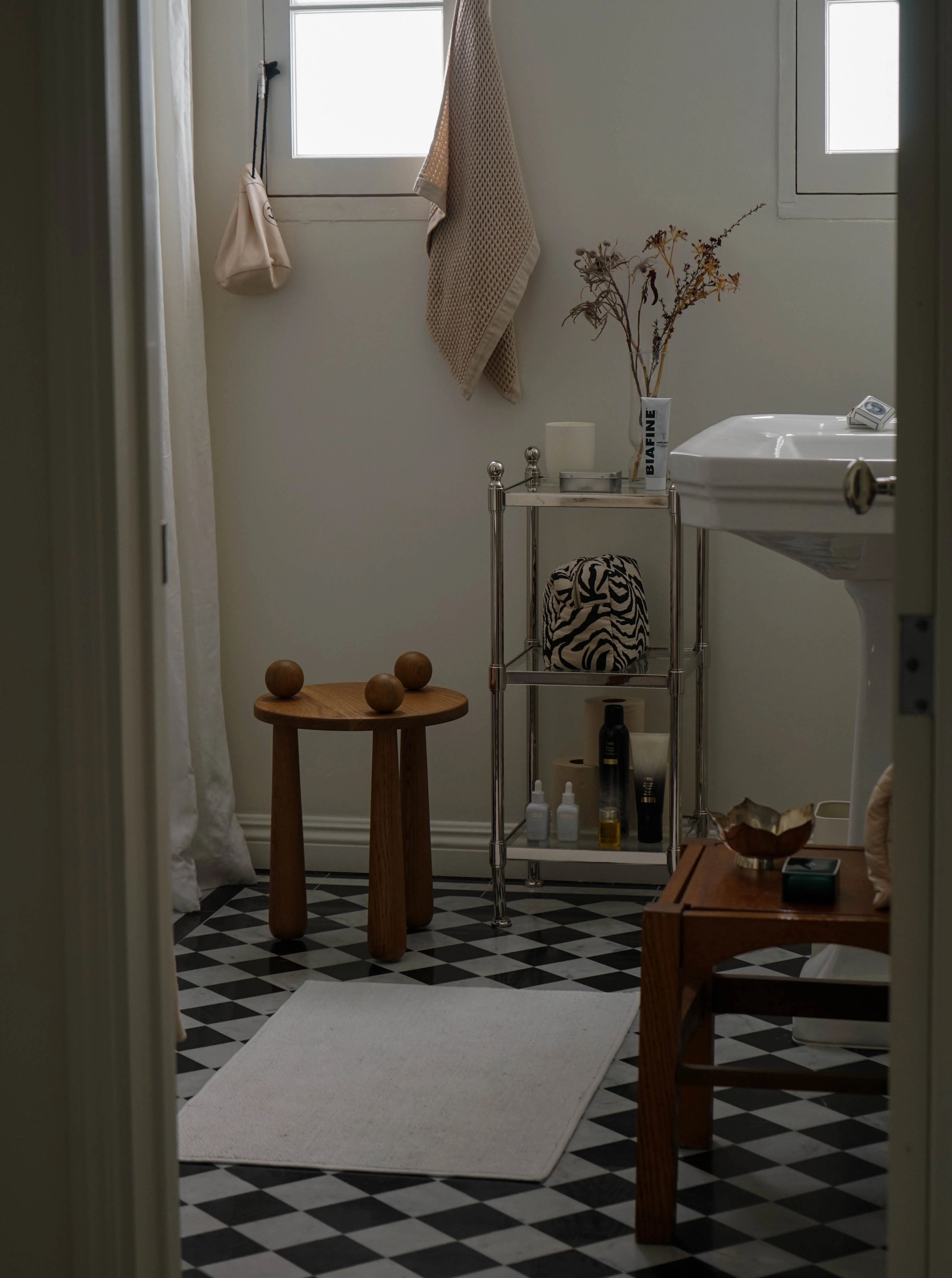 A minimalist bathroom with black and white checkered flooring features a pedestal sink, a TASE GALLERY Dott Stool, Natural, a small table with toiletries, and dried plants in a vase. Two windows with hanging towels provide natural light.