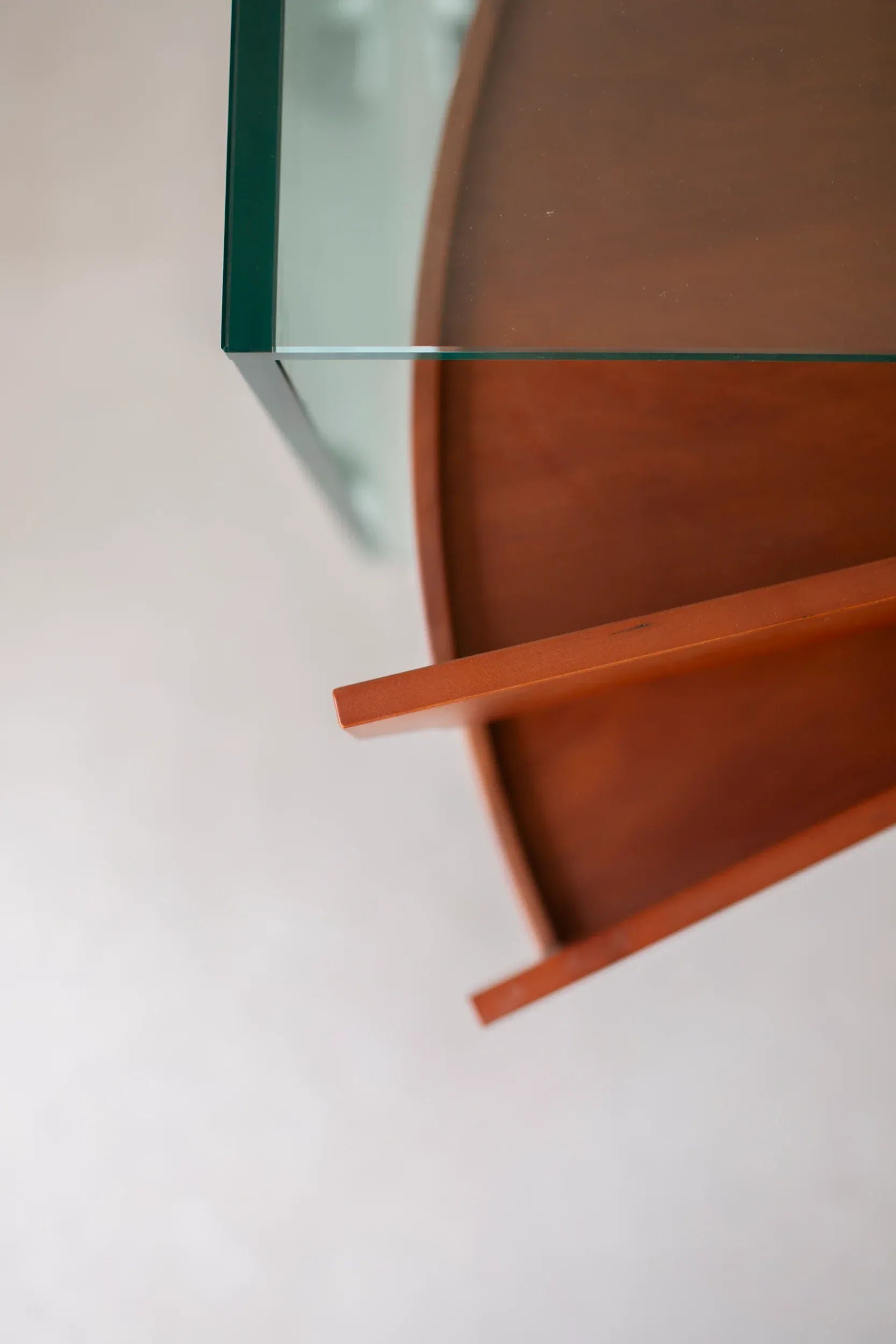 A close-up shot of the Tonelli Camicino Cabinet from CU-RA-TED, designed by Marco Gaudenzi. The cabinet features a tempered glass top with a curved edge, exuding a modern design and minimalist aesthetic. Two wooden support beams are visible beneath the glass top, enhancing its structural integrity.