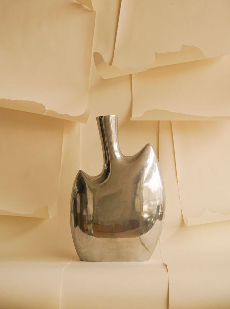 A shiny, polished aluminium Abstract vase XL from Les Objets Amsterdam with a unique broad, asymmetrical shape set against a soft beige background with gently folded paper sheets creating a textured backdrop.