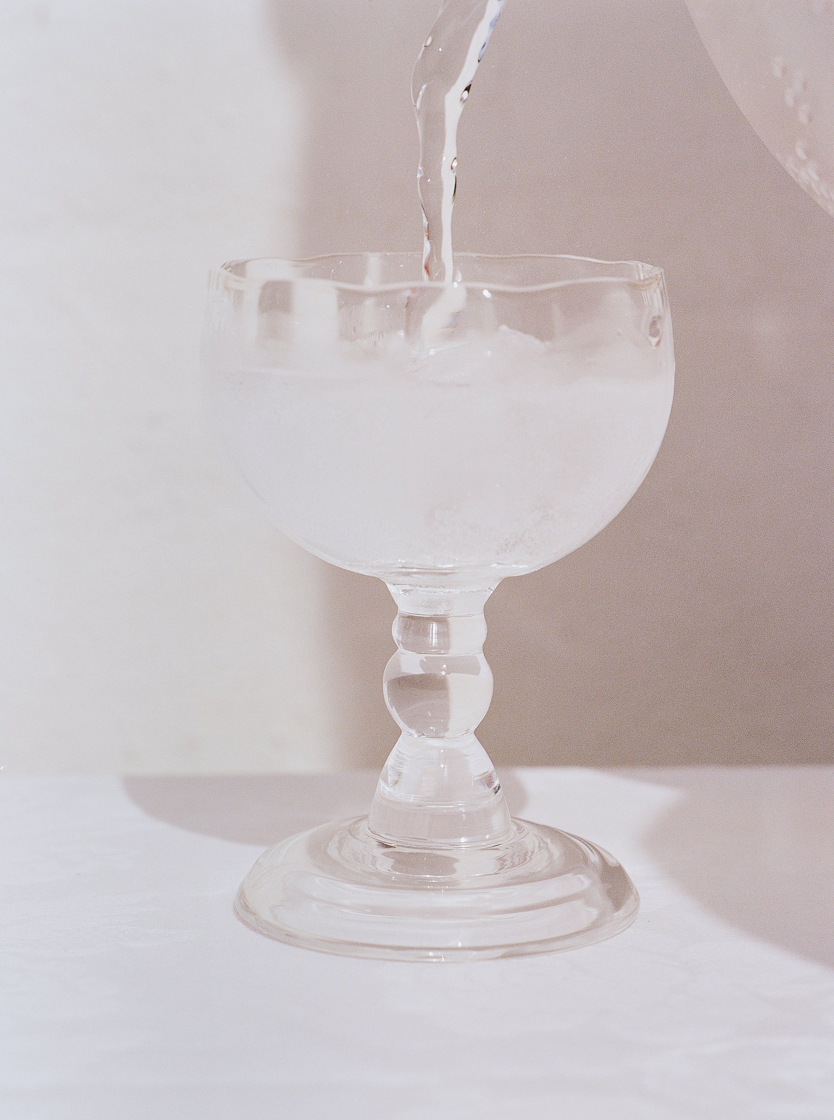 Water being poured into a clear glass Alban Wine Glass from the Akua Objects collection filled with ice cubes, set against a pale background.