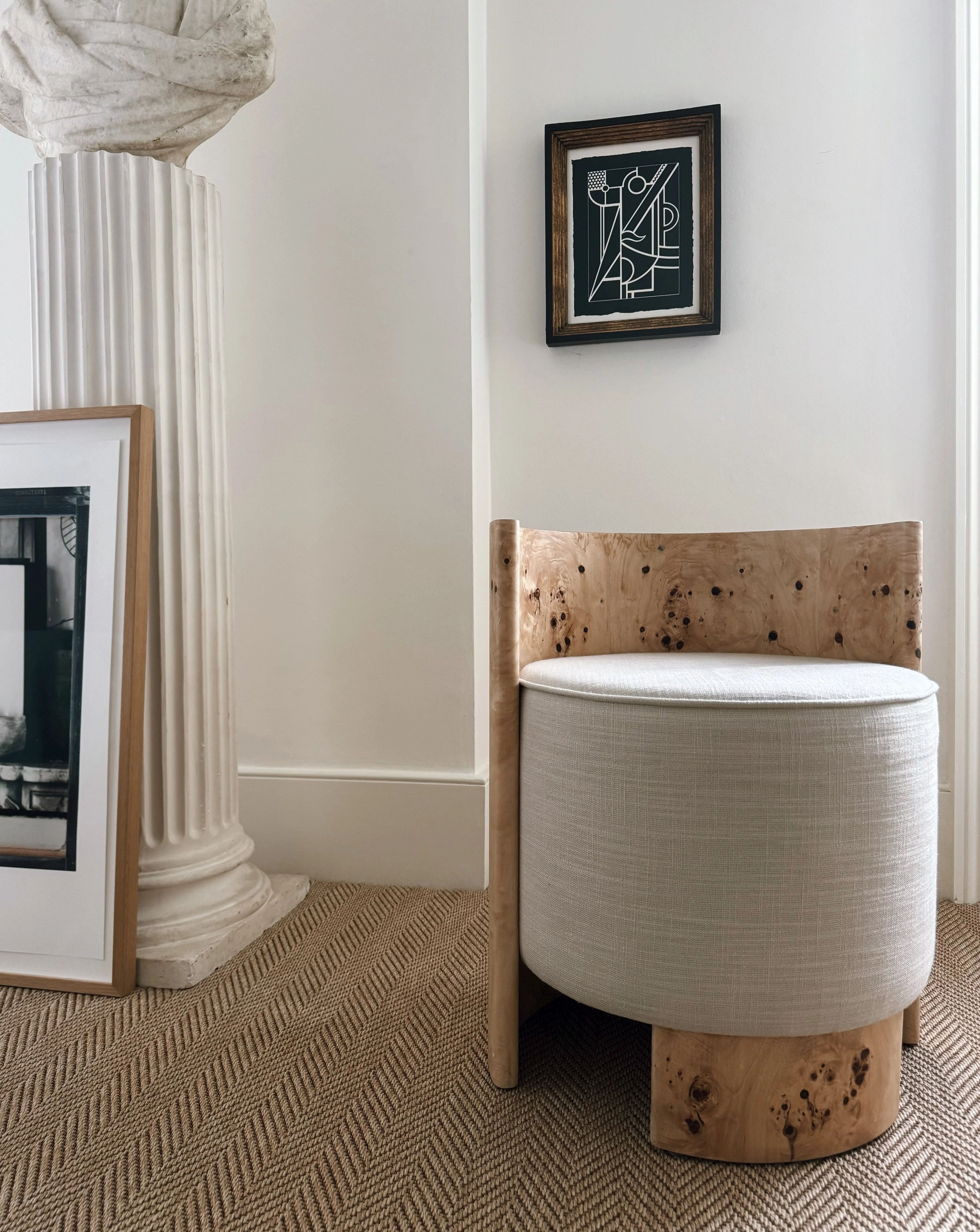 A modern room corner showcases a minimalist seating arrangement featuring the "Livon" Burl Armchair by Ransom & Dunn, with its elegant design reminiscent of ash burl wood. Nearby, a framed black and white photograph leans against a fluted column beside a small geometric artwork, all resting on a textured light brown carpet.