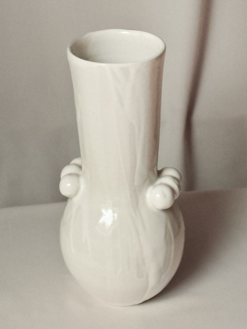 The Vase Solstice I by Landy Rakoto Ceramics is a handmade glossy white ceramic vase with a tall, narrow neck, rounded base, and two decorative side knobs. It's showcased against a simple, light background.