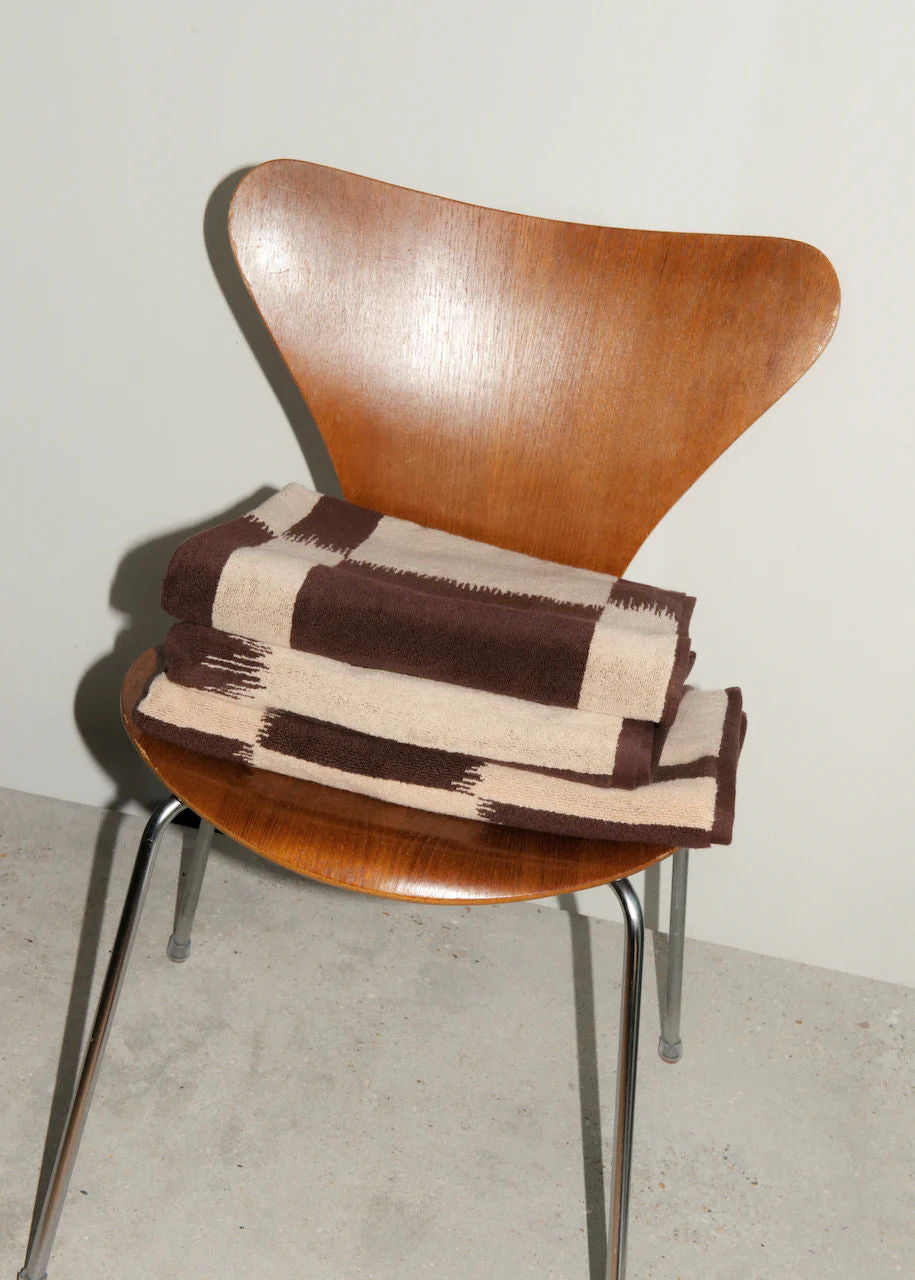 A mid-century modern wooden chair with a curved backrest and metallic legs, holding a folded Autumn Sonata Karin Bath Towel in shades of brown and white, against a neutral background.