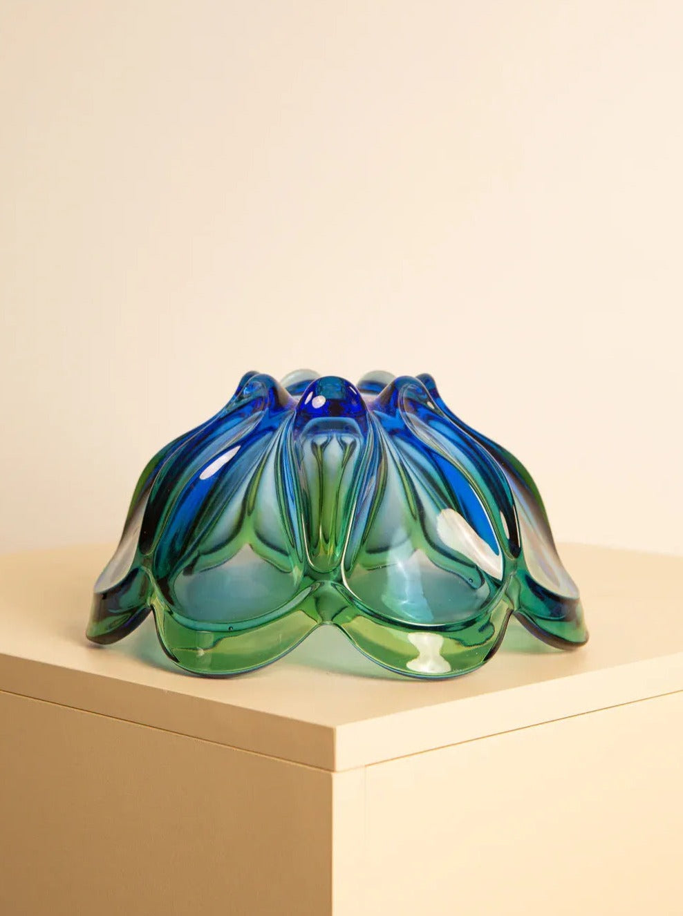 A vibrant, decorative Treaptyque Large vide-poches "Flower" in 60's Murano glass featuring blue and green hues, with an organic, petal-like design. It is displayed on a light-colored rectangular pedestal against a plain, beige background, exemplifying exquisite Italian craftsmanship.