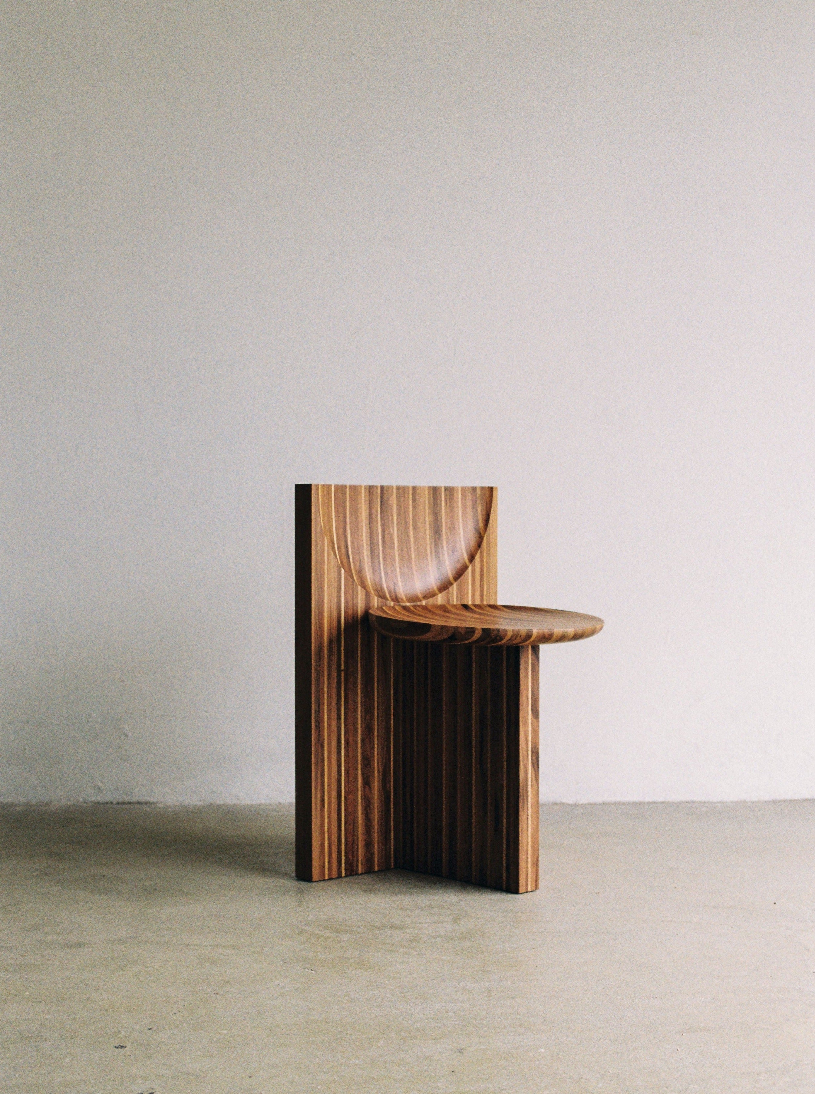 A handcrafted Vaga Chair by Rosana Sousa, made in Portugal from solid walnut and oak wood, stands in front of a plain, light-colored wall. Its unique sculptural design features smooth, curved surfaces and vertical grooves, creating a minimalist yet striking appearance. The floor has a plain surface with a slightly rough texture.