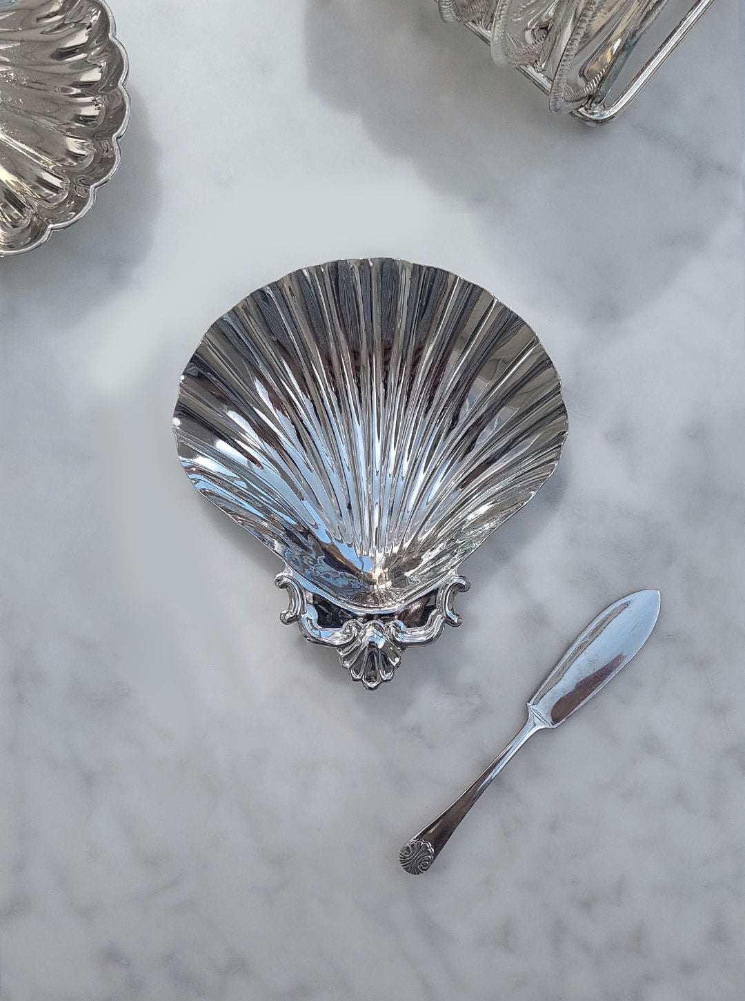 A Shell Butter Dish by Les Objoies glistens on a white marble surface, complemented by an assorted knife with an ornate handle.