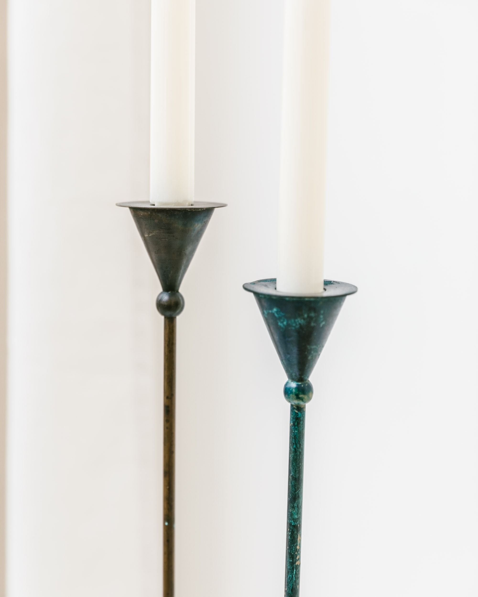 The Set of 2 Candleholders by Bottega Jacobs exudes a Postmodern vintage aesthetic from the 1980s. Crafted with slender metal forms and taper candles, one holder displays a slightly darker and rustic aged patina. Their minimalist design and conical shape are accentuated against a softly blurred background.