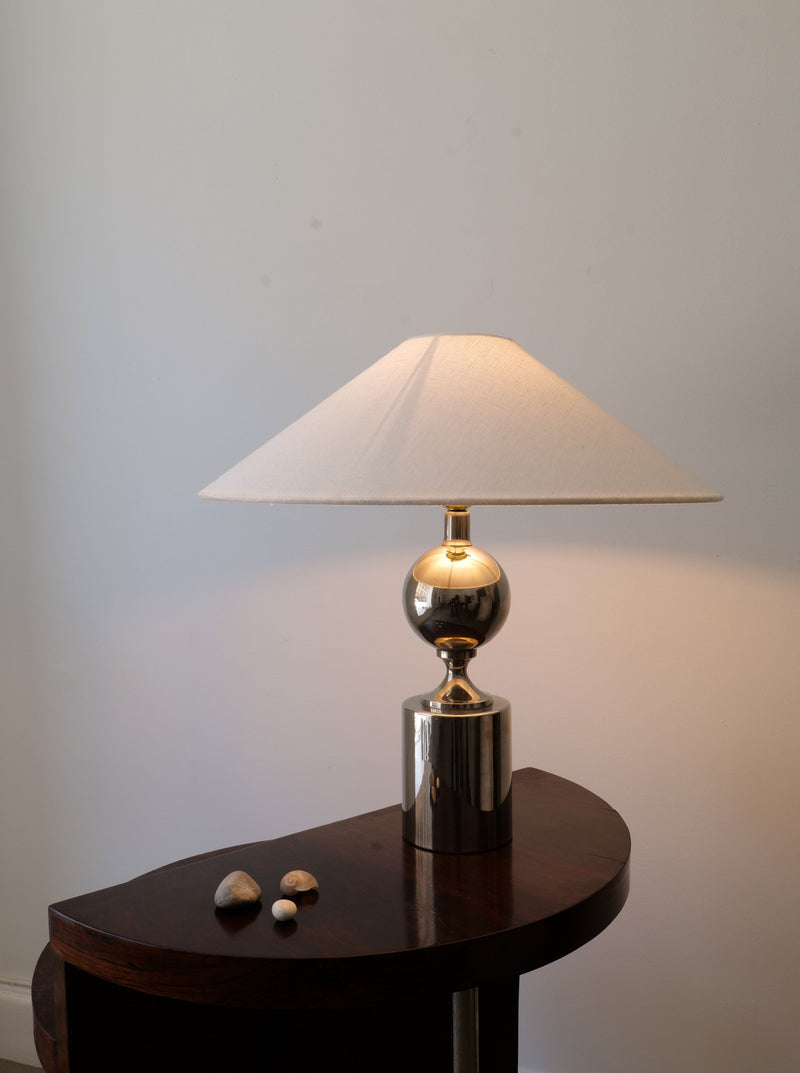 A modern "Barbier Lamp 1970s" by Collection apart with a beige lampshade is placed on a dark wooden side table. The table also has three small stones, scattered to the left of the lamp. The background features a plain white wall.