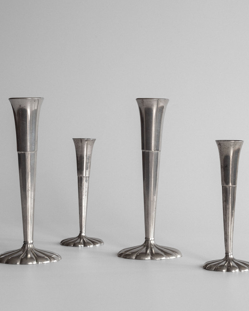 A set of four sleek, metallic Spigel vases, varying in height and featuring elongated designs, stand on a plain surface with a neutral background. Reminiscent of Scandinavian style and echoing Just Andersen's Art Deco influence, they boast modern minimalism with reflective surfaces.