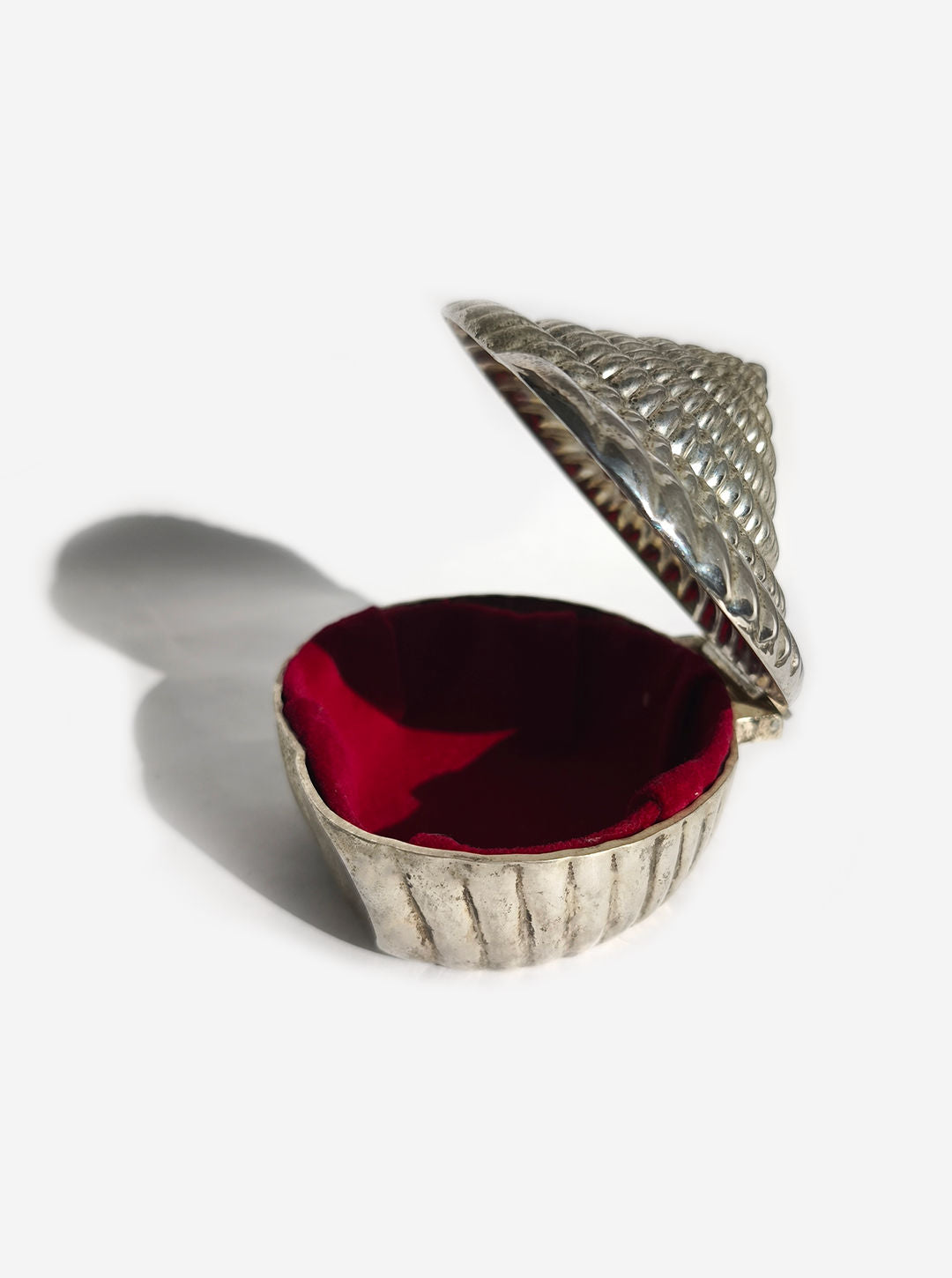 The Les Objoies Silver-plated Shell Box, charmingly shaped like a spiral seashell, boasts a luxurious red velvet lining. Slightly open, it casts a delicate shadow on the white background, inviting you to admire its exquisite interior.