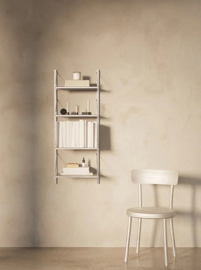 A minimalist room features a light-colored wall-mounted Shelf Library Single Section | Stainless Steel | H1084 / W40 by FRAMA with neatly arranged books, decorative items, and a candle. Below the modular system is a simple white wooden chair on a polished floor. The scene is bathed in soft, natural light.