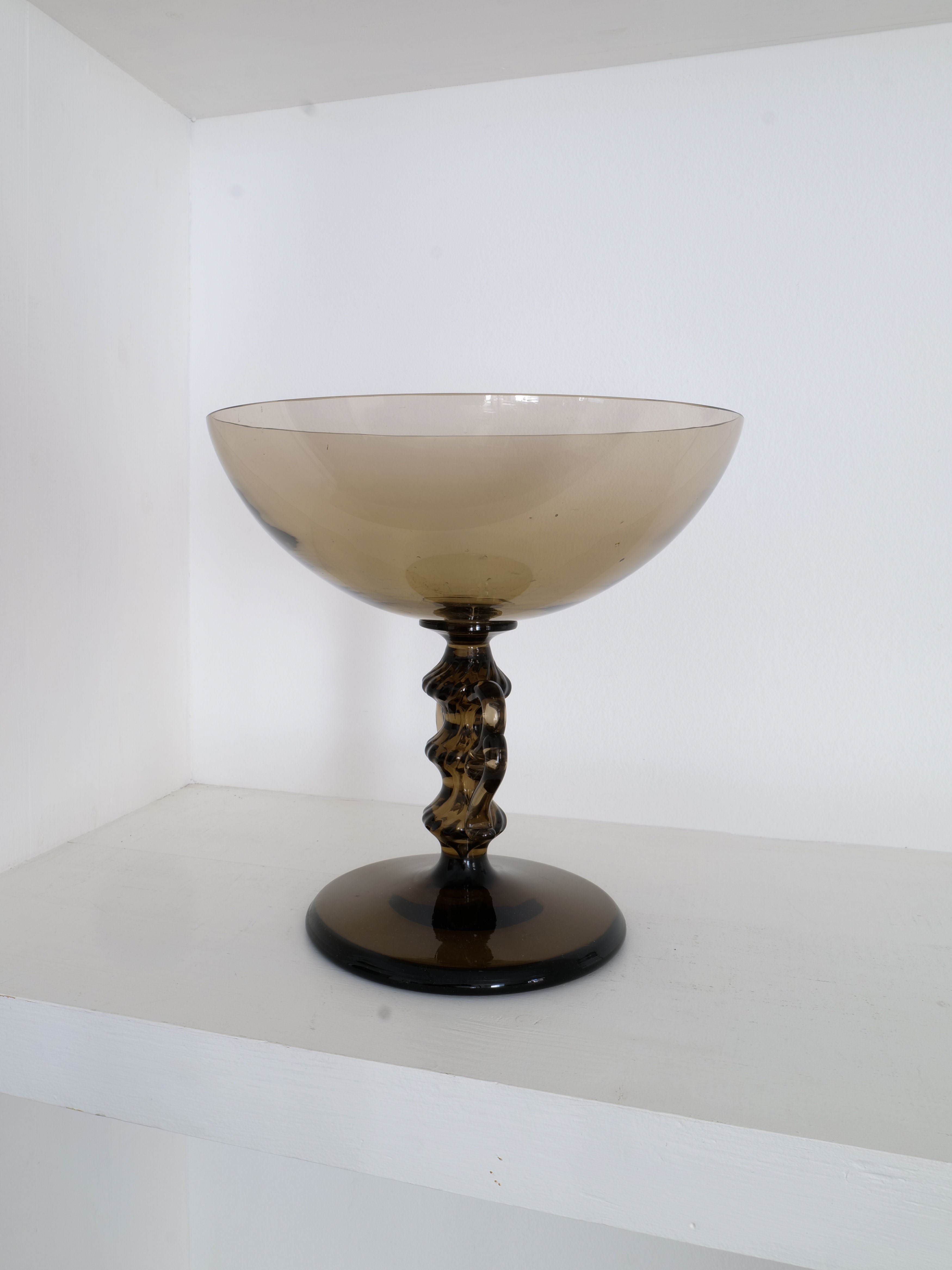 A translucent, smoky gray Glass Bowl Simon Gate designed by Simon Gate for the Collection apart is displayed on a white shelf. The collectible piece features a wide, shallow bowl, a decorative stem, and a solid round base, all in the same smoky gray color against a plain white wall.