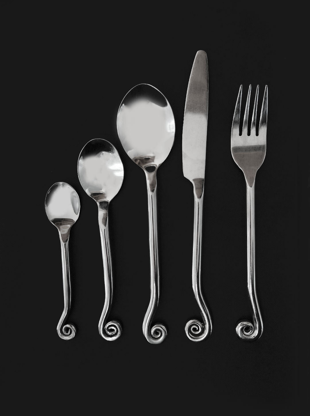 The Les Objoies Set of 5 Swirling Cutlery displays five shiny silver utensils—small spoon, larger spoon, soup spoon, knife, and fork—on a black background. Each handle ends with an exquisite spiral design.