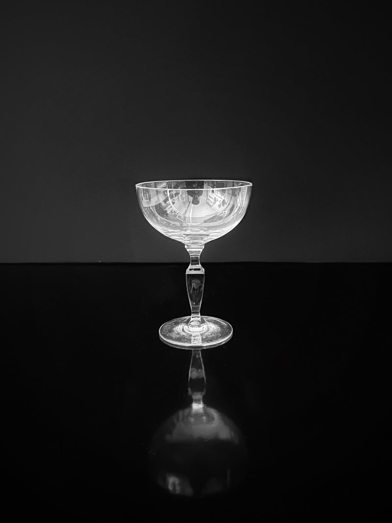 A dazzling champagne coupe from the "6 Mid Century Crystal Champagne Coupes" by Dodo Vintage sits on a reflective black surface against a dark background, casting a subtle reflection below.