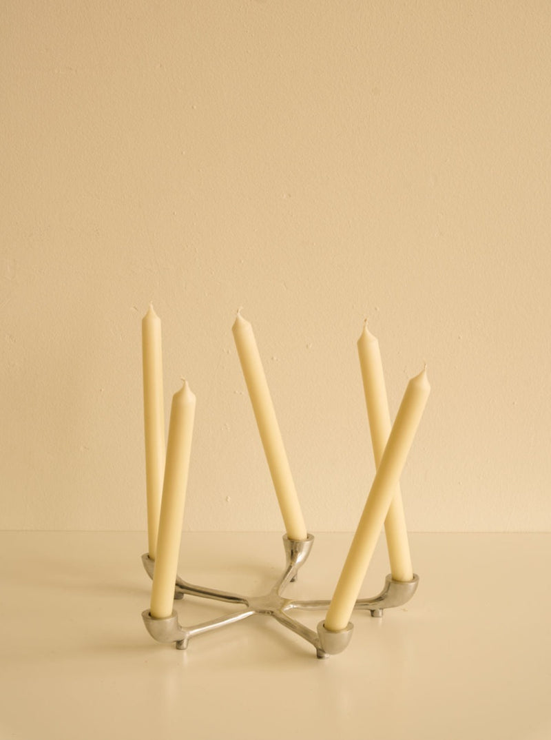Five beige candles of varying heights arranged in a Les Objets Amsterdam sleek candelabra against a soft beige background.