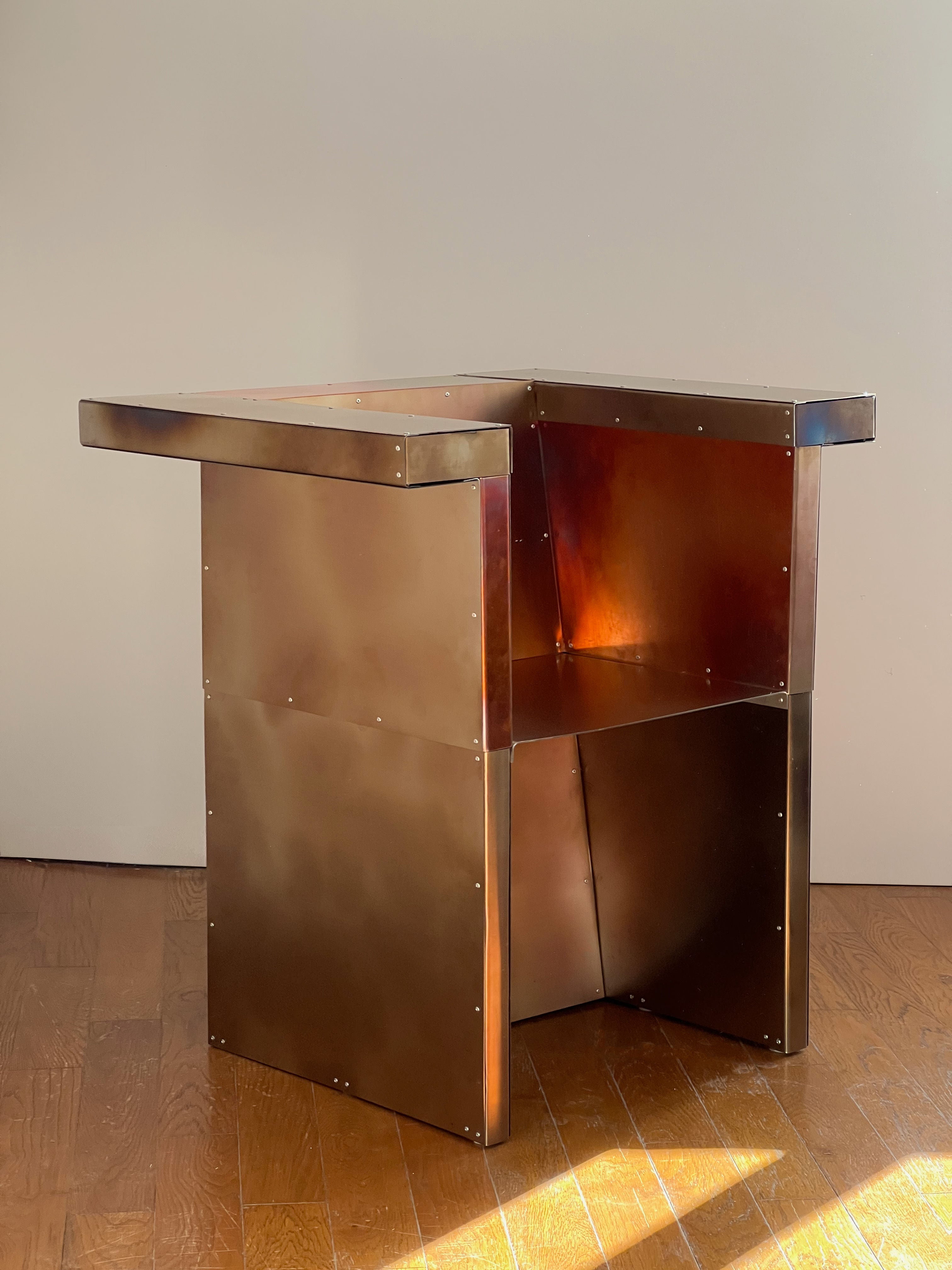 The Throne chair by Theo Galliakis features an angular, modern design with visible bolts and reflective metallic panels, crafted from industrial materials. It stands on a wooden floor against a plain light-colored wall.