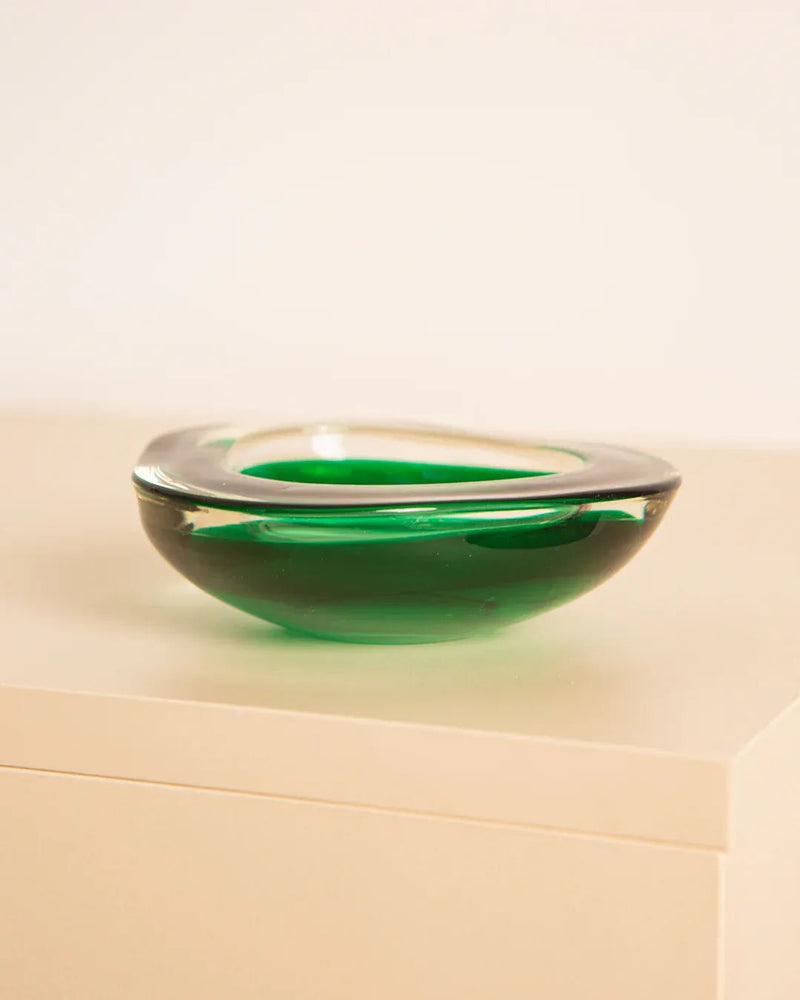 The Treaptyque Vide-poches "Triangle" Murano 60's, a small green Murano glass bowl with a clear rim, rests elegantly on a light-colored surface against a plain background, showcasing the artistry of Italian glassmaking.