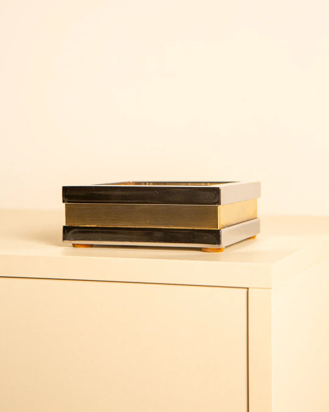 A Treaptyque Italian Silver-plated Metal and Brass Box from the 60s, with a glossy black and gold finish, elegantly rests on a light surface against a plain background. It features two stacked layers with gold accents, showcasing exquisite craftsmanship.