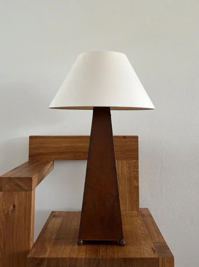 A CU-RA-TED Obelisk Table Lamp with a white conical lampshade and a triangular wooden base sits on a wooden table against a plain white wall. This stylish lamp's design features clean, minimalist lines, exemplifying refined sophistication while complementing the natural wood tones of the room.