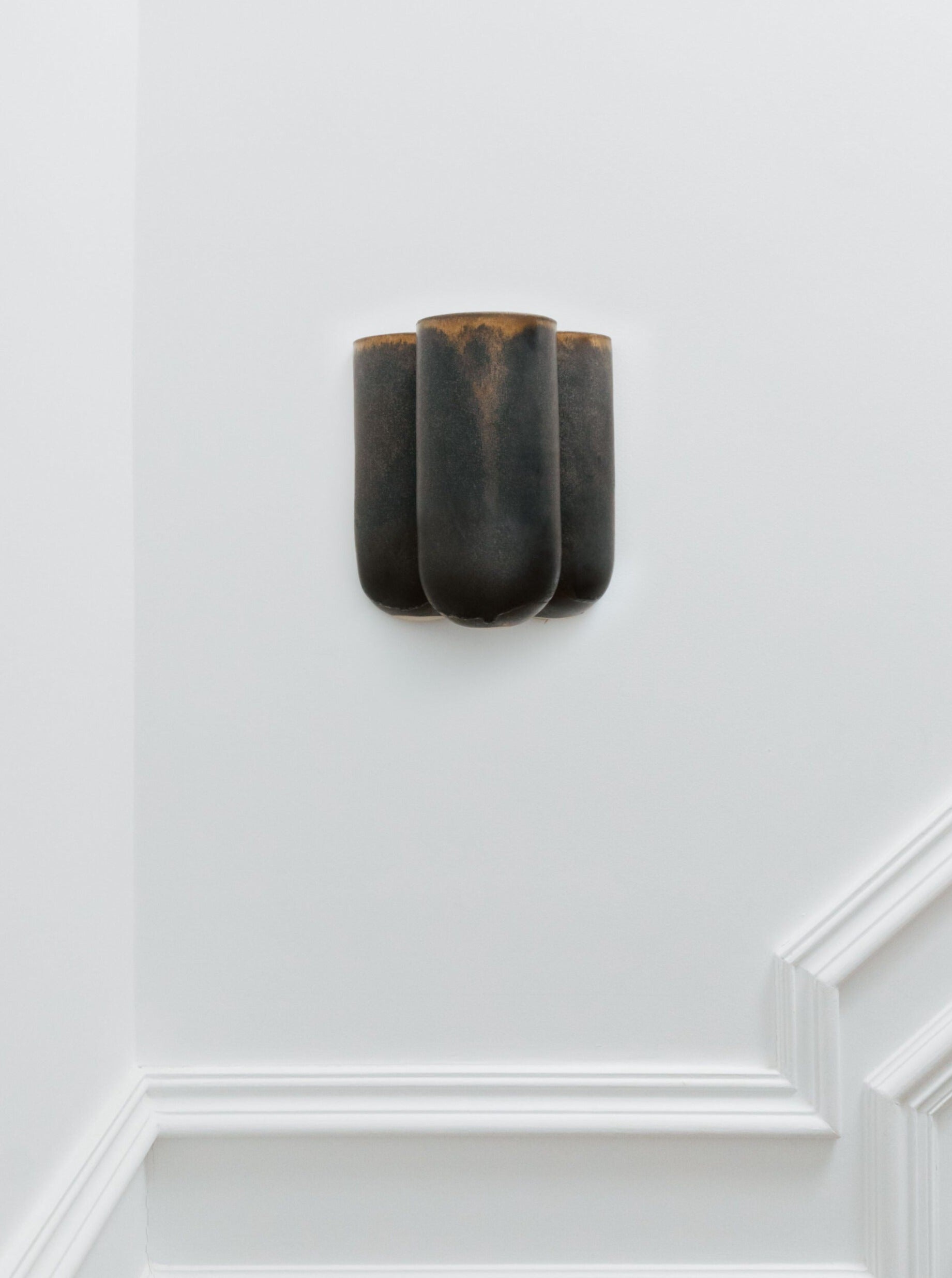 The Lisa Allegra Wall Lamp MOOR PLUS BRILLANCE, a black ceramic sconce with a smooth, curved sculptural design, is installed on a white wall with decorative molding. The sconce holds multiple bulbs and features an enamel finish that enhances its modern, minimalist aesthetic.