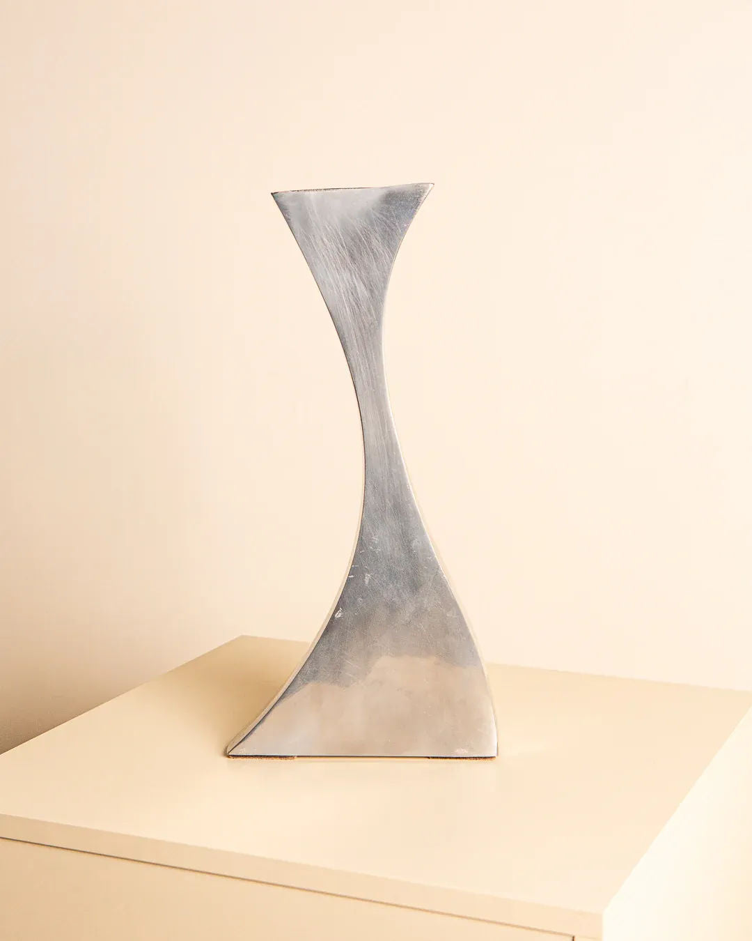 A minimalist sculpture from Treaptyque, the Curved Aluminum Vase 80's, exudes modernism and elegance with its twisted metallic design as it stands on a light-colored square pedestal against a plain off-white background.