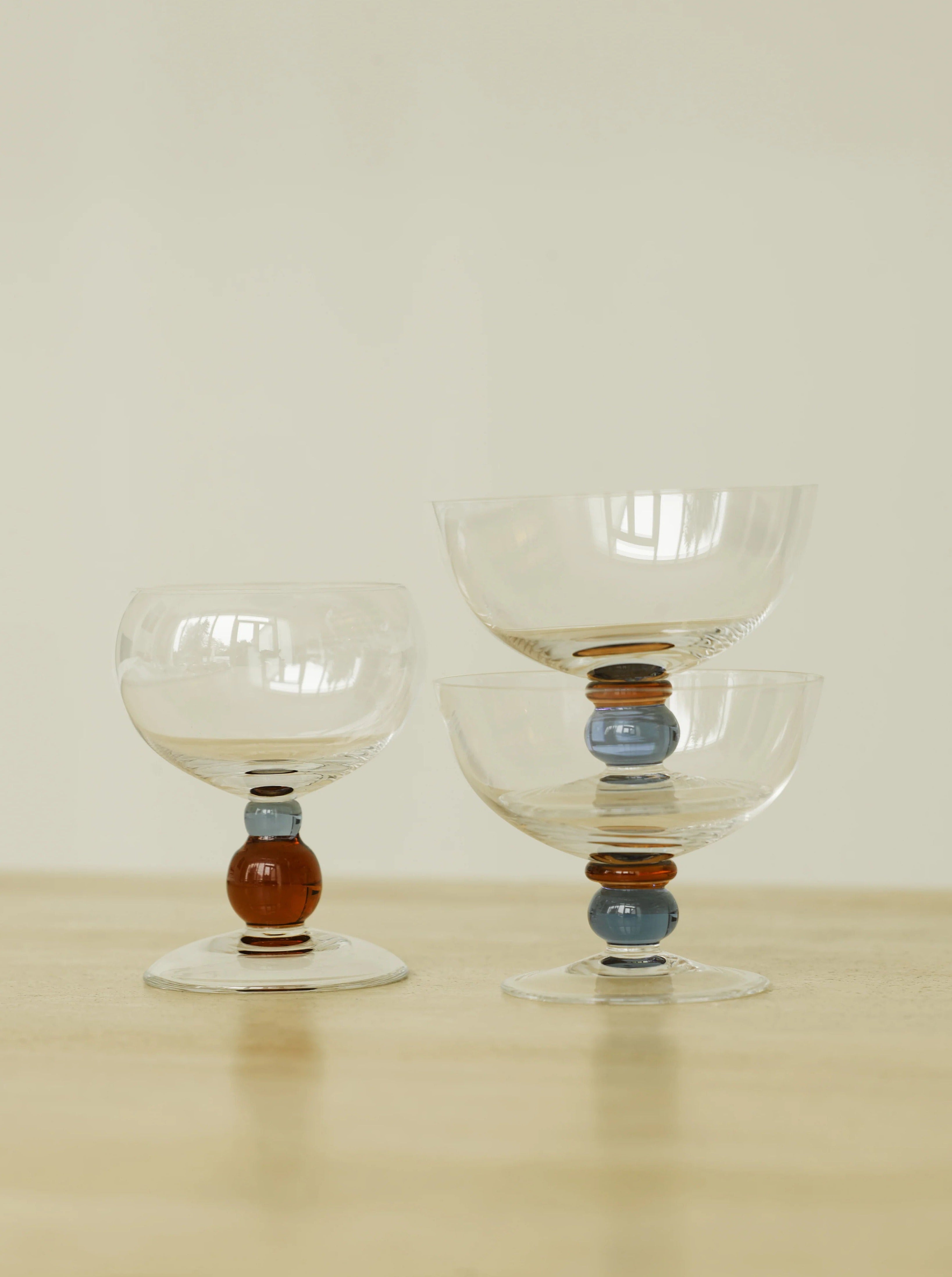 Three elegant Noam Wine Glasses - Set of Four by Akua Objects with colorful, round decorative elements on their stems are arranged on a plain light-colored surface. The goblets vary in size, with two stacked on the right and one standing alone on the left. The background is neutral and unobtrusive.