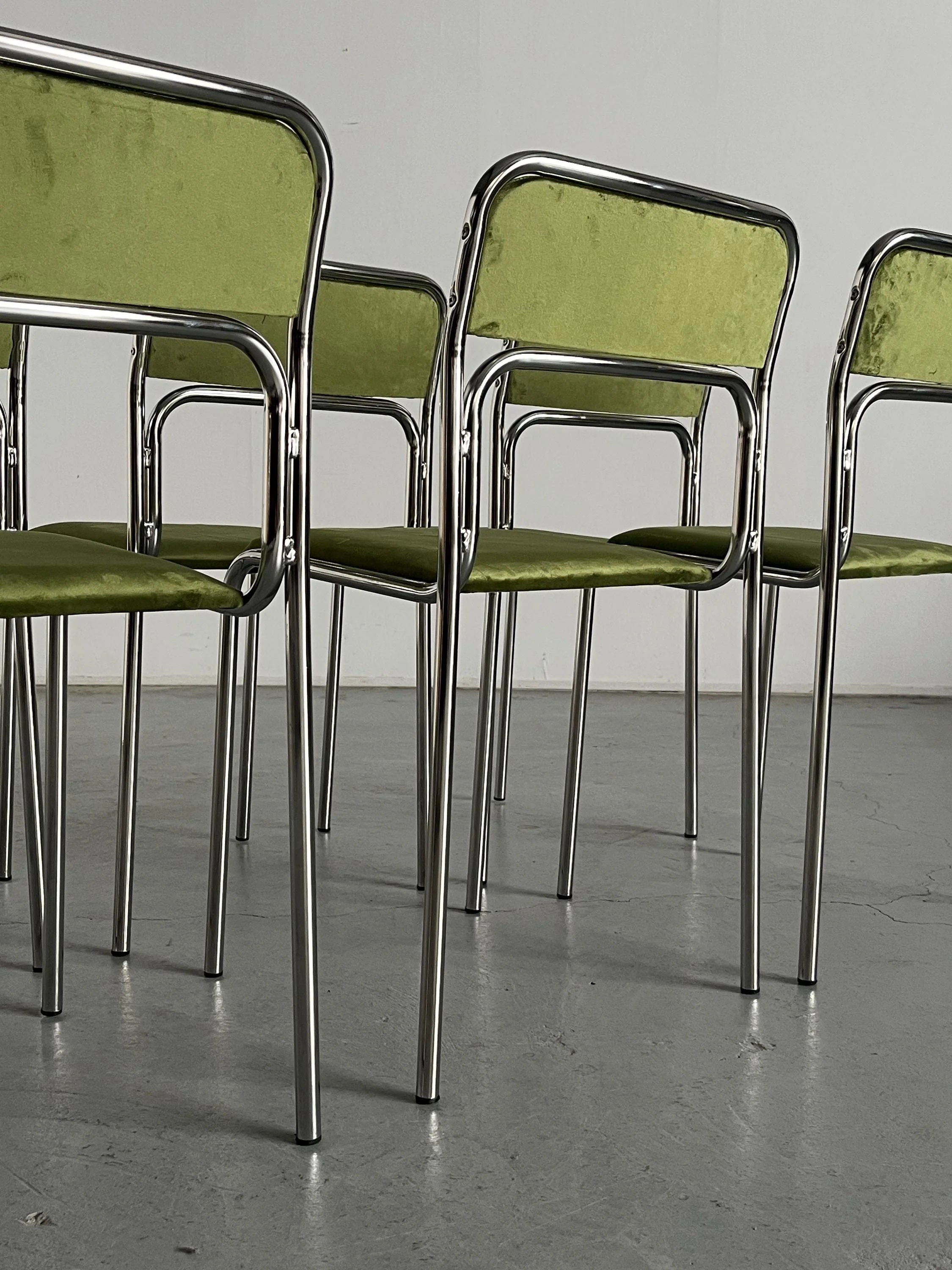 A Set of 6 Bauhaus Chrome and Green Velvet Chairs by Cherry Cargo are neatly arranged on a gray floor, featuring sleek tubular chrome frames with green upholstered seats and backs, in an empty room with a plain white wall.