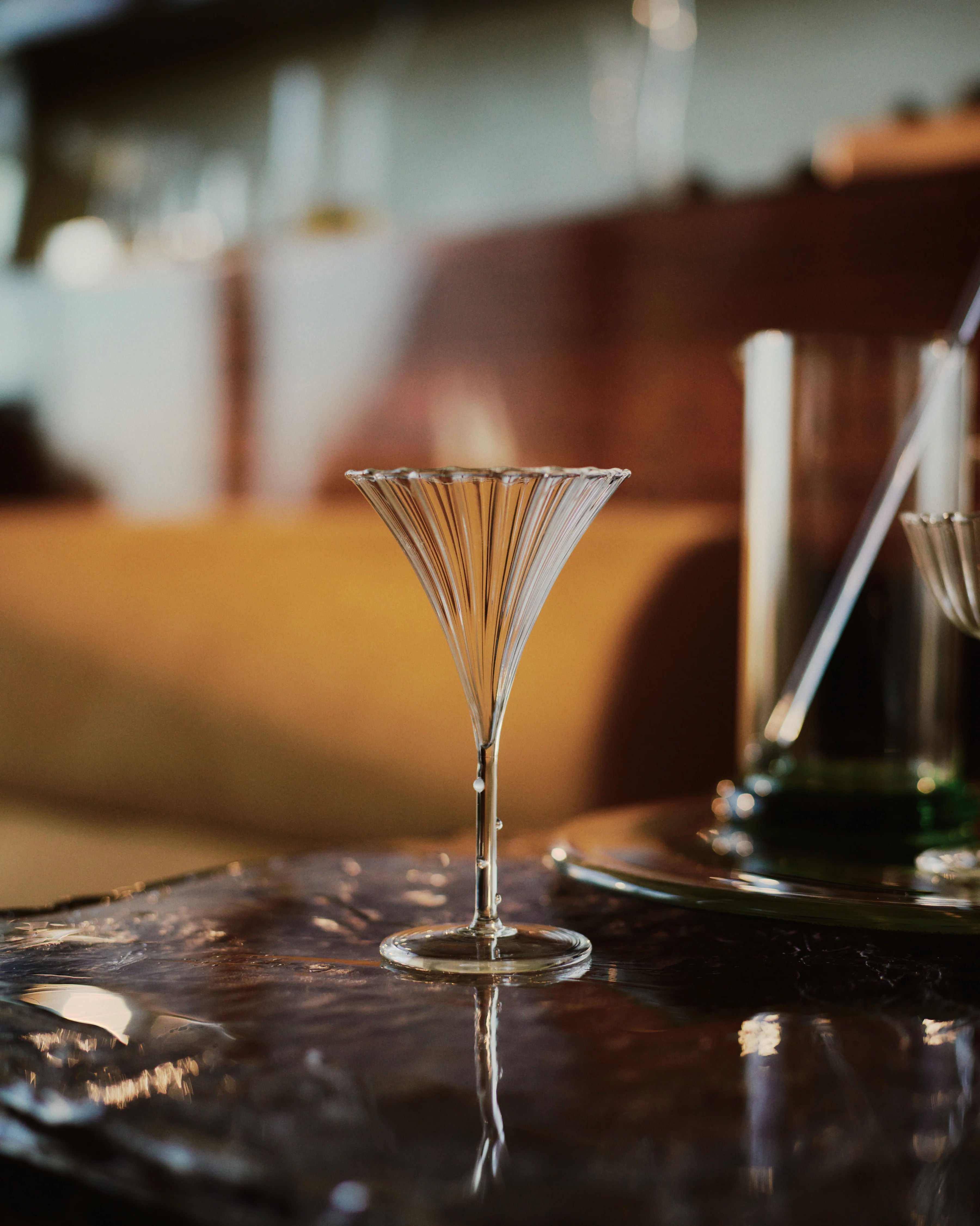 A delicate Trumpet Aperitif Glass - Set of Four by Sophie Lou Jacobsen with vertical etched lines sits on a reflective surface. In the background, blurred objects and a warm, ambient setting suggest a home or cozy bar environment. The focus is on the glass, highlighting its intricate design. This set of 4 glasses enhances any collection.