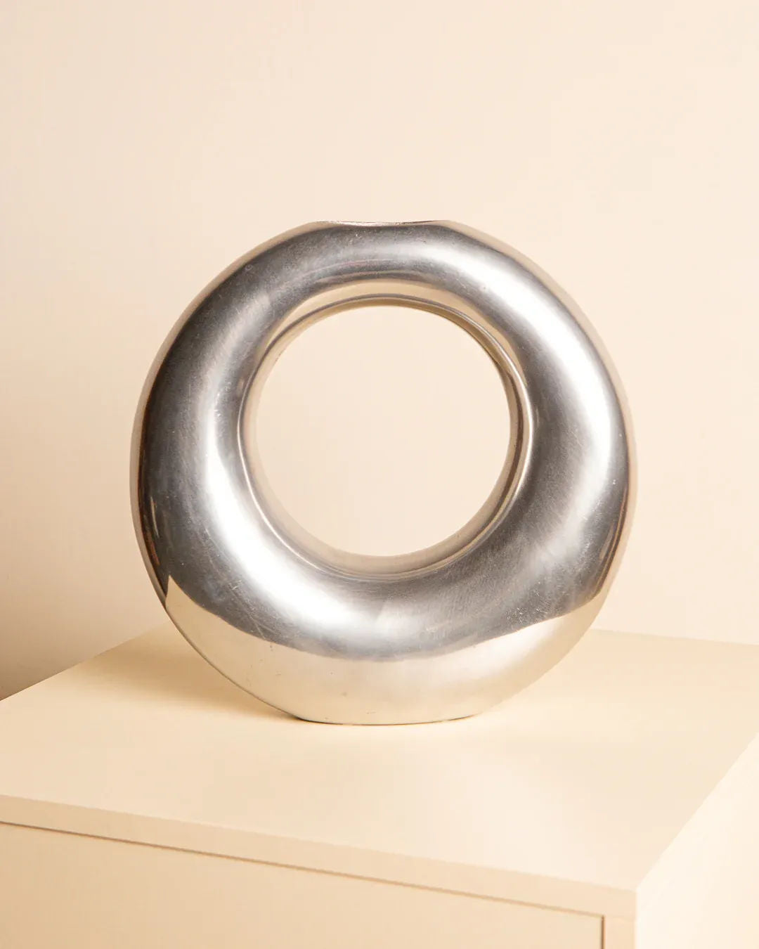 A modern, shiny silver circular sculpture named the Aluminum “Sac” Vase by Treaptyque, featuring a hollow center, is displayed on a light-colored wooden surface against a beige background.