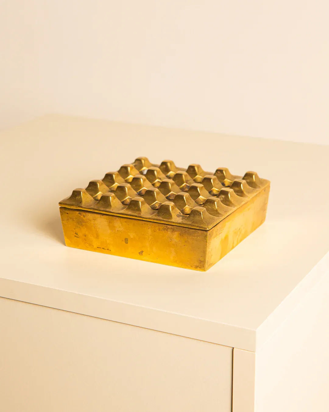 A brass ashtray by Holger Bäckström from Treaptyque, square in shape with a golden hue and textured geometric pattern, rests on a white surface against a plain background.