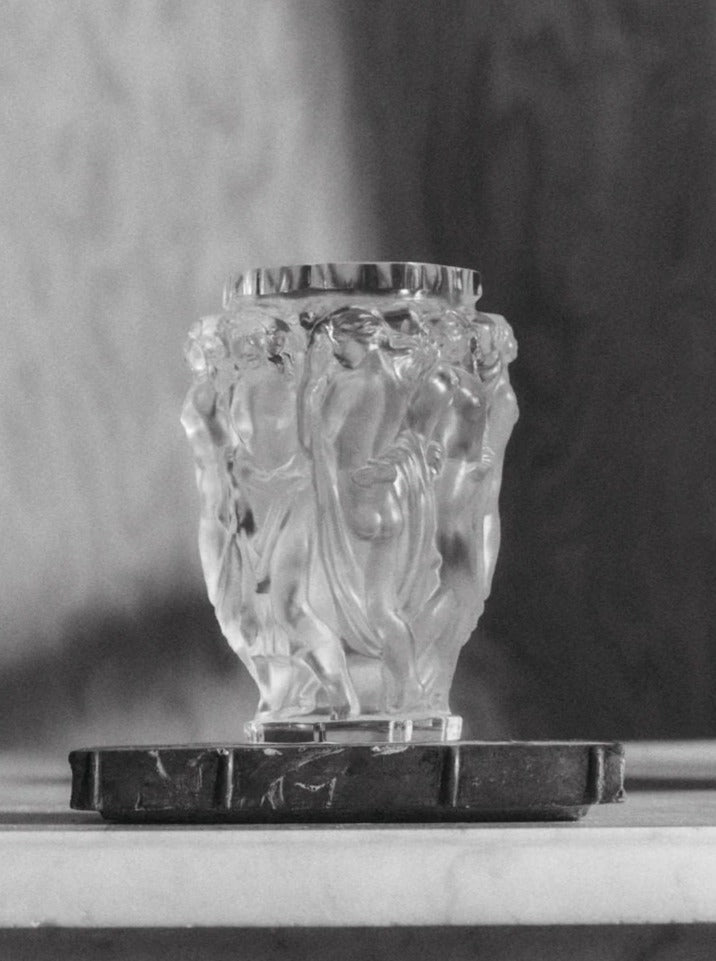 A black-and-white photograph depicts a small ornate Michael Vase by Akua Objects with intricate carvings of human figures draped in flowing garments. The vase is placed atop a stone base; light and shadow create highlights and depth, bringing out the details in the glasswork, sure to captivate antique collectors.