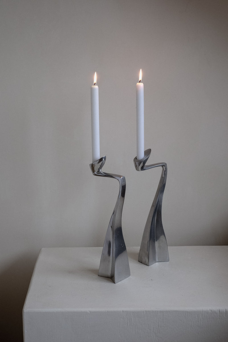 Two lit white candles rest on Pair of Candleholders by Matthew Hilton 80's from septembre studios. They sit atop a plain white surface against a neutral wall, showcasing their sleek, contemporary design with smooth curves, ideal for collectors.