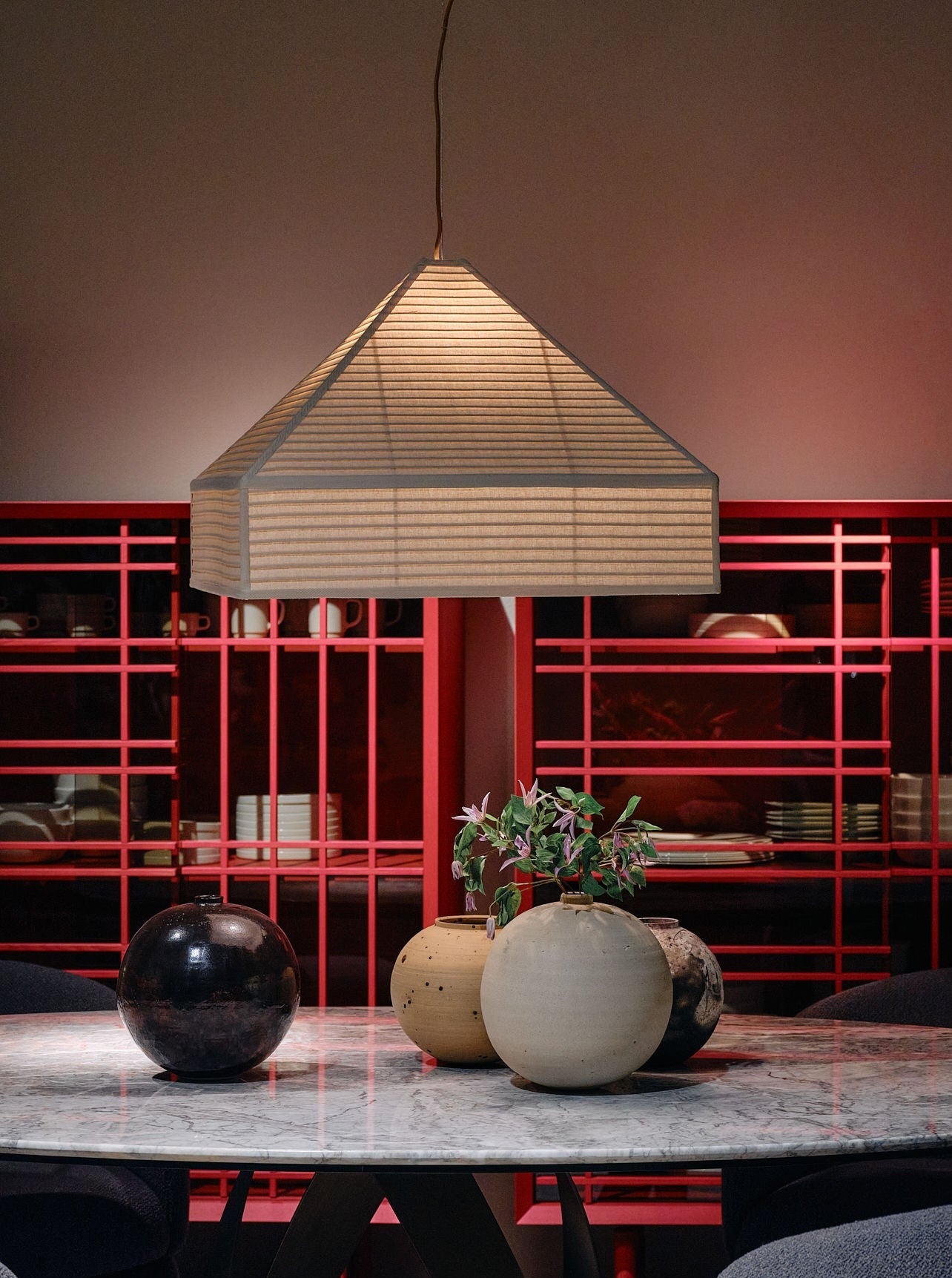 A stylish dining area featuring a circular marble table adorned with three spherical vases, one containing a small plant. The backdrop is a striking red shelving unit holding various dishware, and overhead is the Pyramid Hanging Lamp by Nassi Lamps. Cozy gray chairs surround the table.