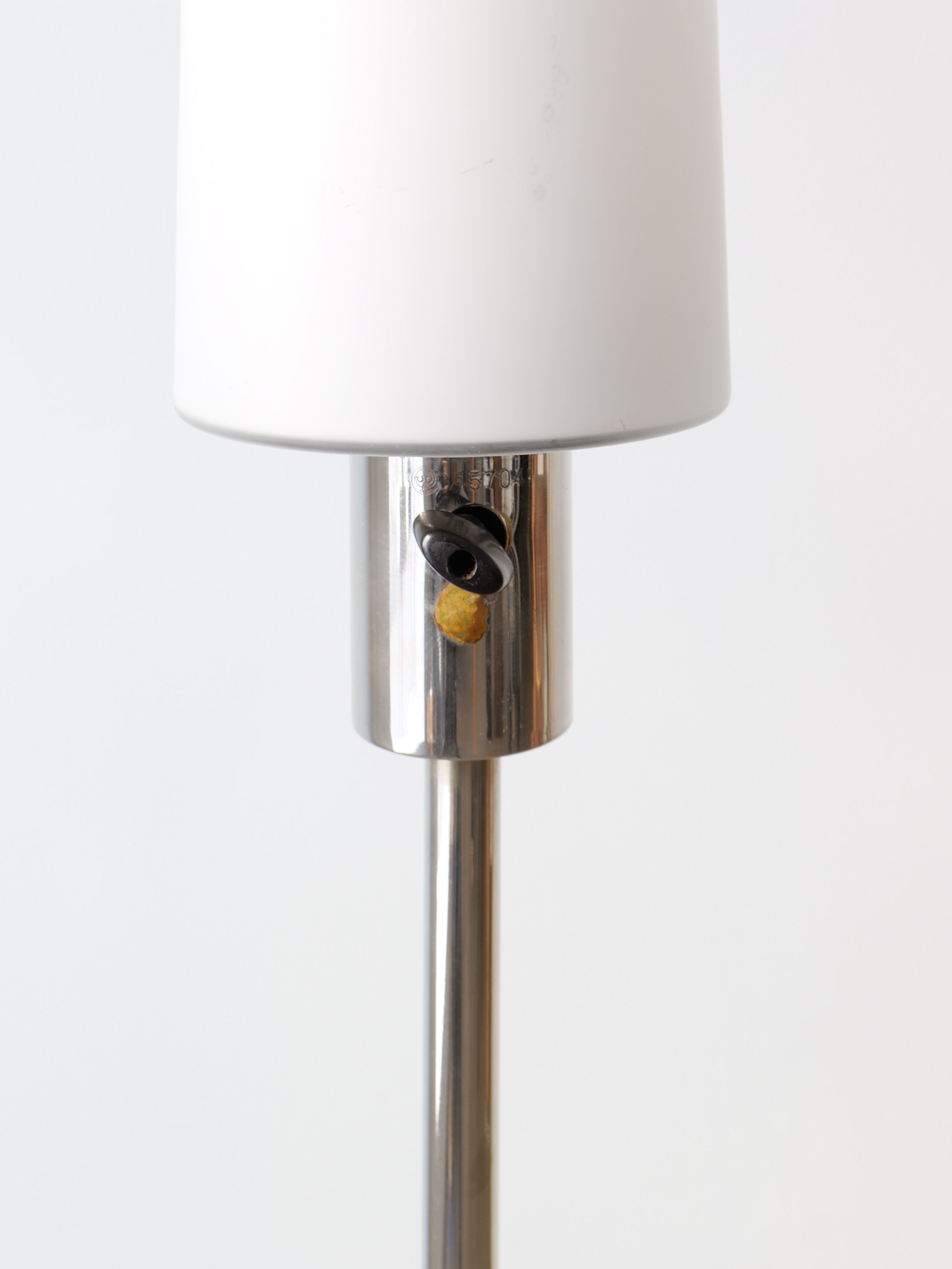 A close-up view of a modern metallic Deco Floor Lamp Harald Notini 1940s from Collection apart with a white lampshade. The lamp's switch is visible on the top part of the metallic base, featuring a small yellow detail beneath it. The background is plain and white, keeping the focus on the lamp's Swedish Modern design inspired by Arvid Böhlmarks Lamp Factory.