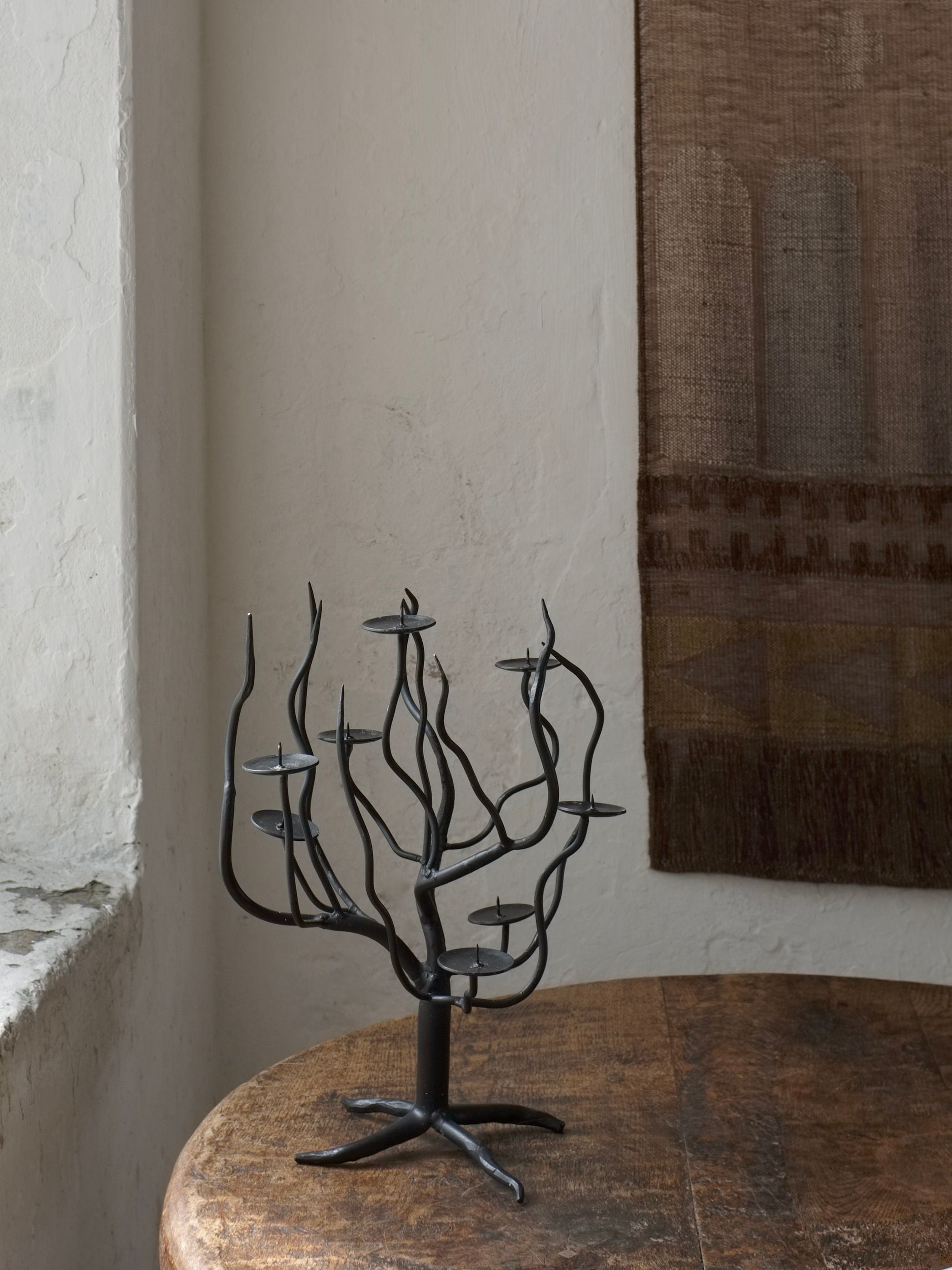 A Hand-forged Metal Sculptural Candle Holder by Veter Vintage, featuring branches reminiscent of a tree and finished in black metal, is placed on a wooden table beside a rustic white wall with a tapestry in the background. This sculptural piece, inspired by 1960s design, has empty candle holders at the tips of its branches.