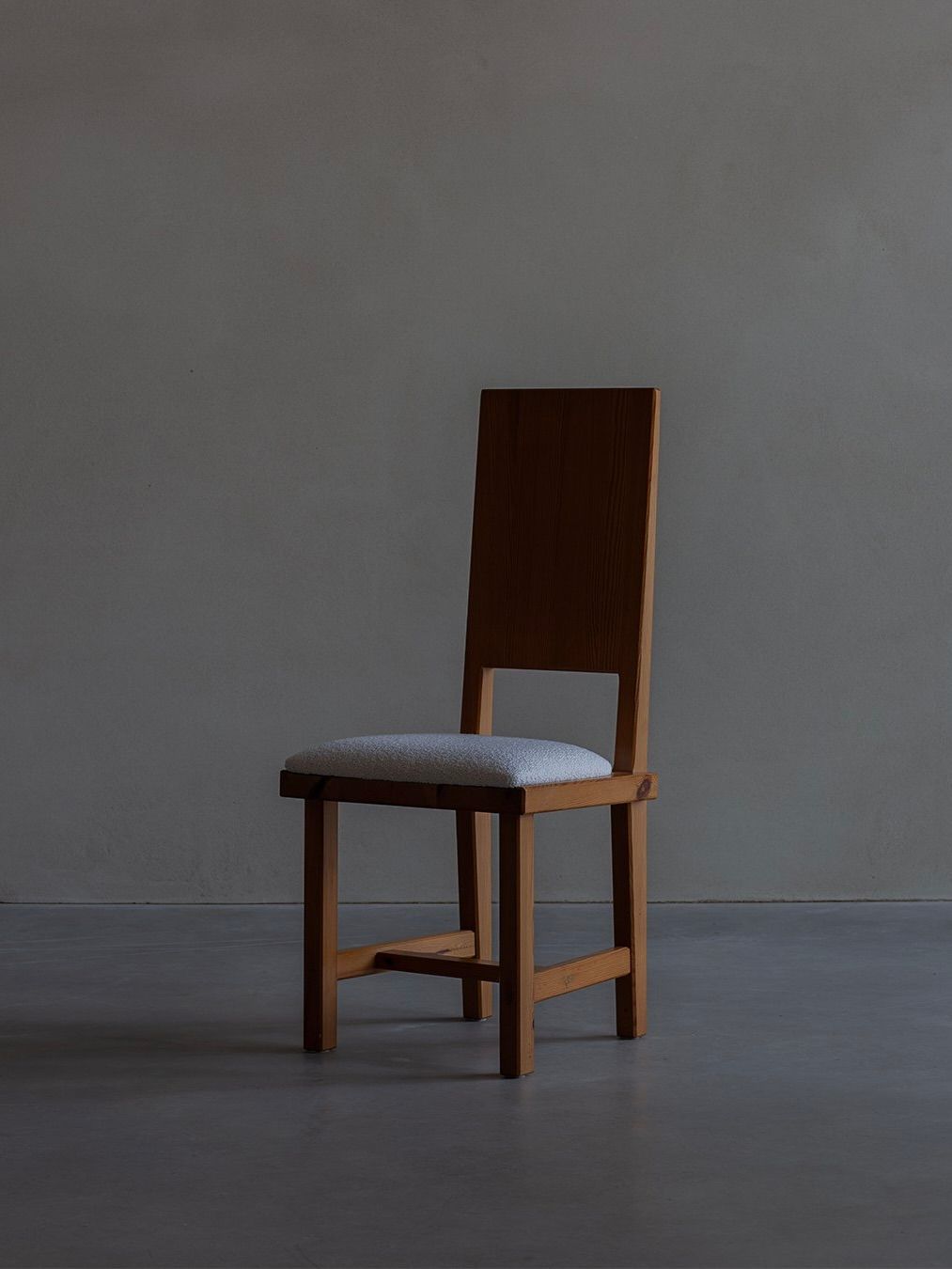 In a dimly lit room with a gray background, the Bicci de' Medici "Göran Malmvall - Model 553" wooden chair features a tall backrest and cushioned seat, exuding Swedish-inspired minimalist design and capturing Göran Malmvall's craftsmanship.