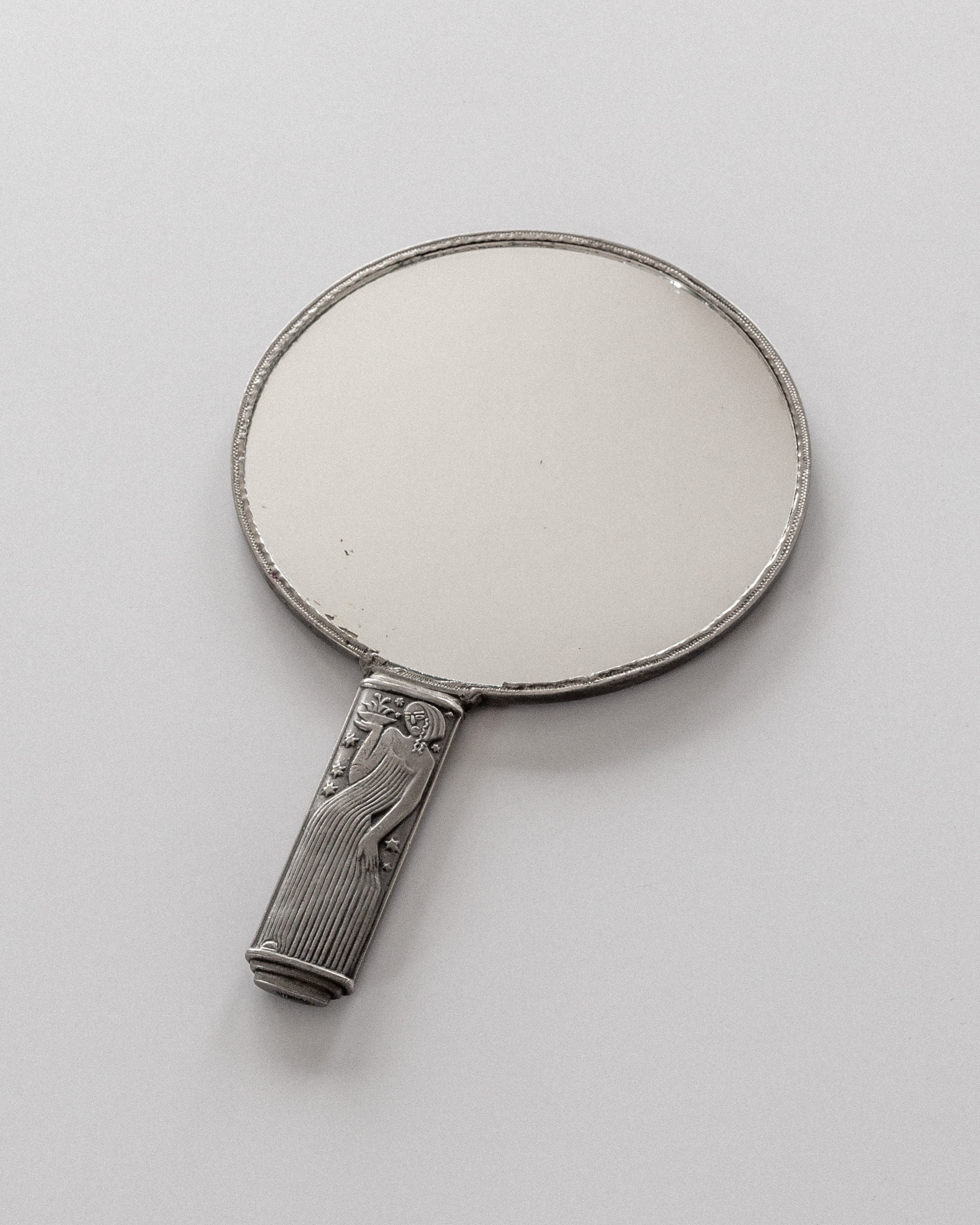 A vintage pewter hand mirror with an ornate handle and engravings, featuring Spigel's Modernist Metal Mirror by Sylvia Stave from the 1930s, rests on a light gray surface. The oval-shaped mirror showcases a metallic finish reminiscent of modernist design.