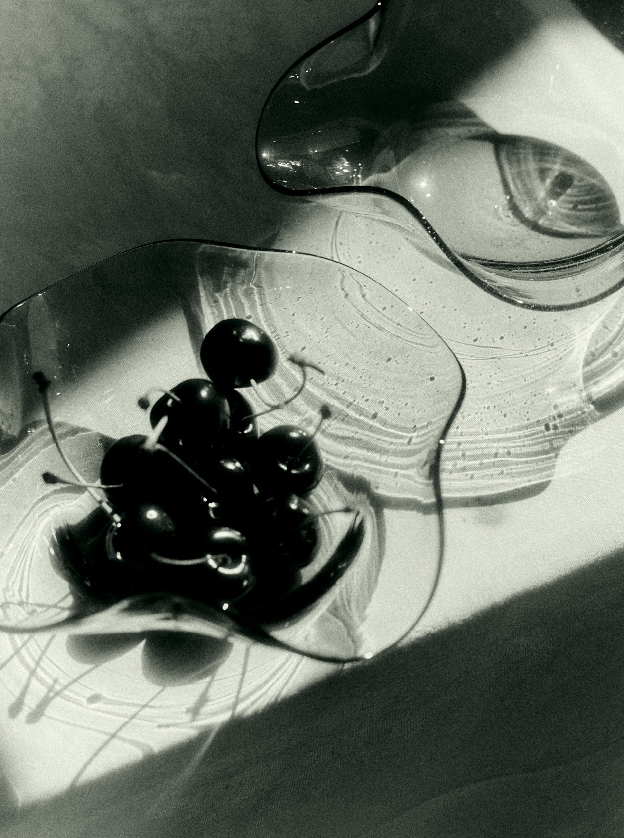 A black and white photograph featuring two exquisite bowls of handmade Venetian glassware. The closer bowl, an Akua Objects Michele Bowl Small, contains a bunch of cherries with stems attached, while the farther Akua Objects Michele Bowl Small is empty. Both bowls cast intricate shadows on the white surface beneath them, highlighting their craftsmanship.